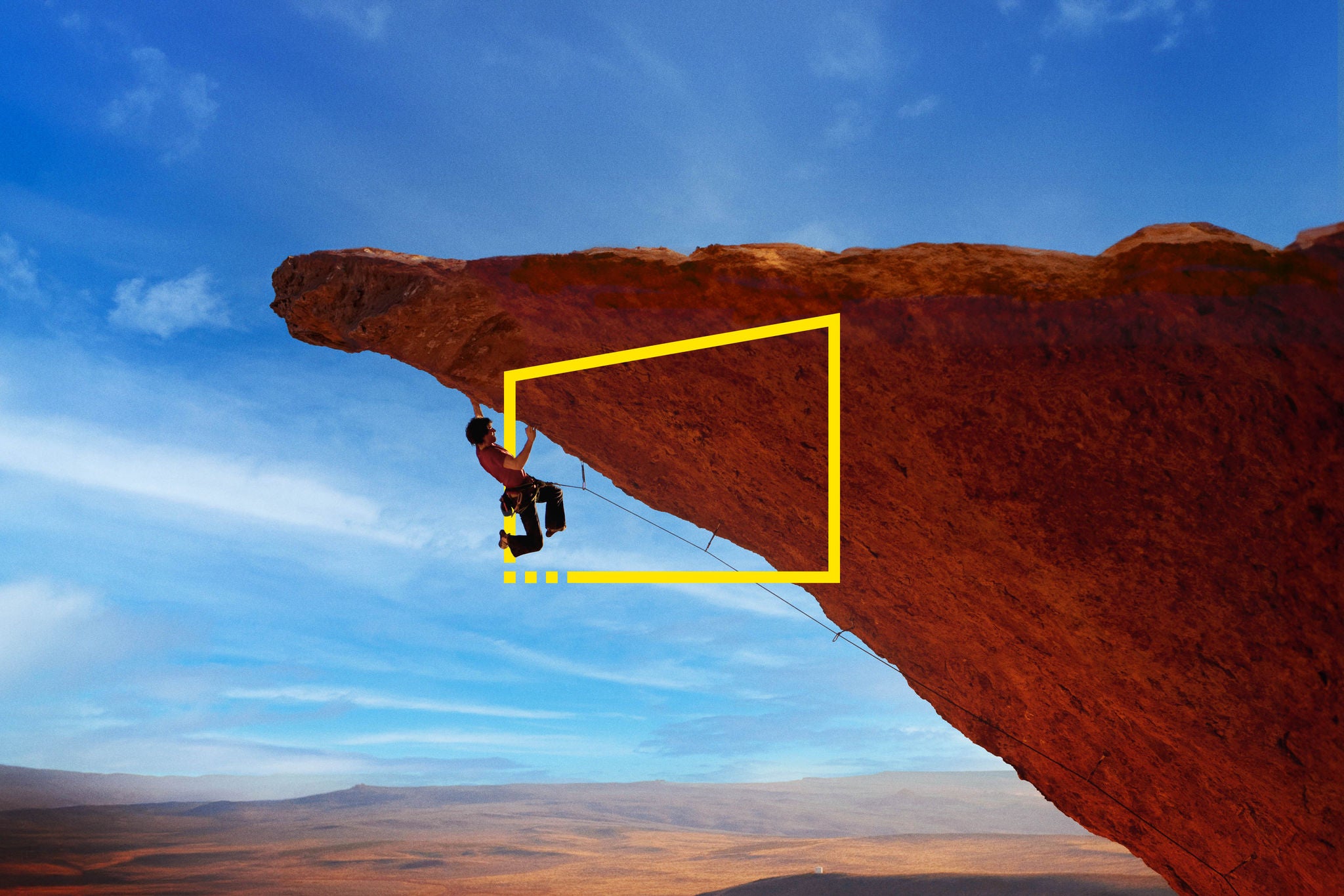 Professional rock climber Joe Kinder lunging for a hand hold on a steep over hanging rock wall while both feet are peeled away from he rock due to the difficulty of the move in South West Utah. He is high above ground and the rolling hills of the desert can be seen behind him and off in the distance.