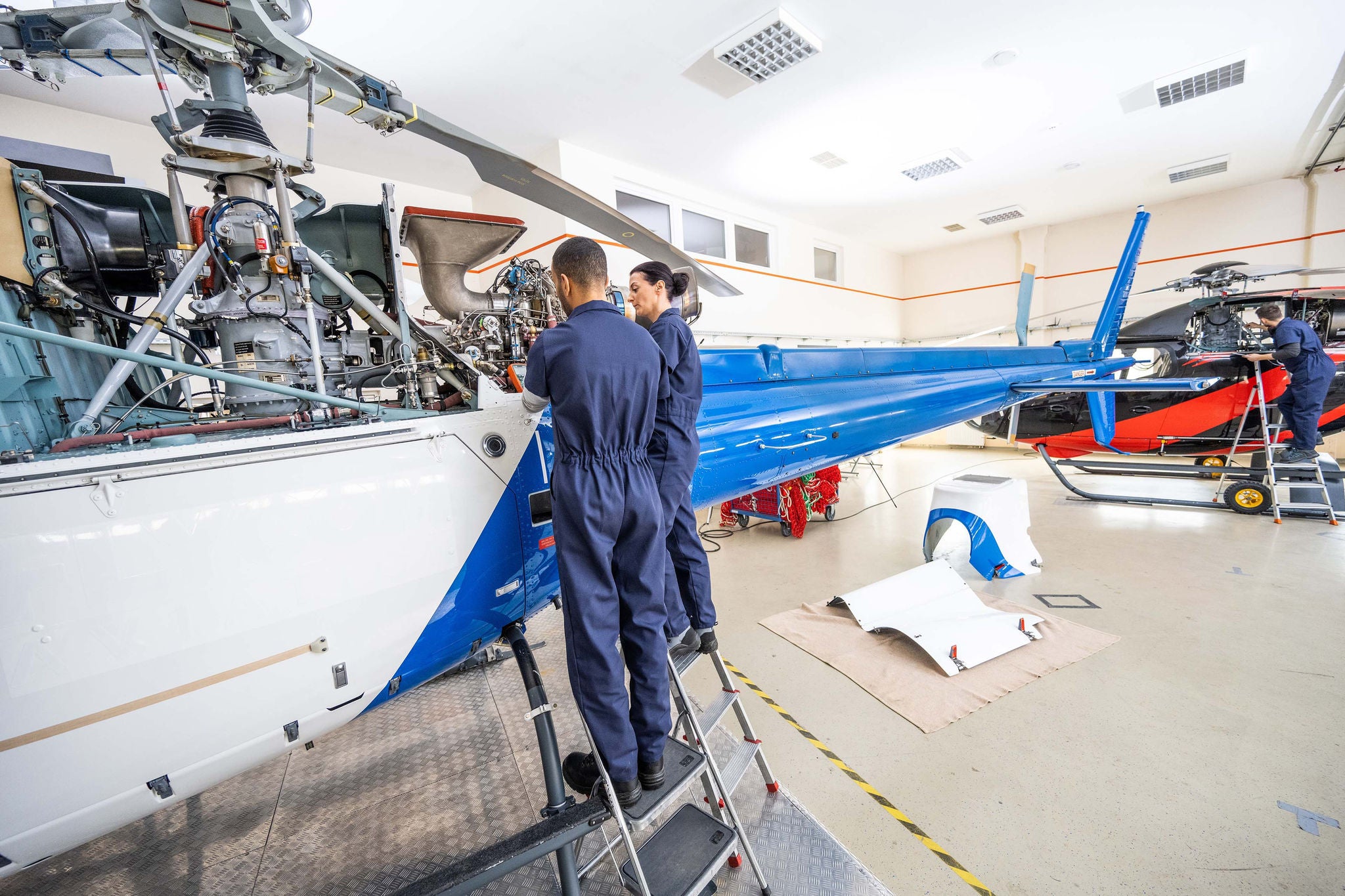 Two airplane mechanics fixing helicopter engine, back view. Male and female coworkers standing on ladder and repairing jet, long angle shot