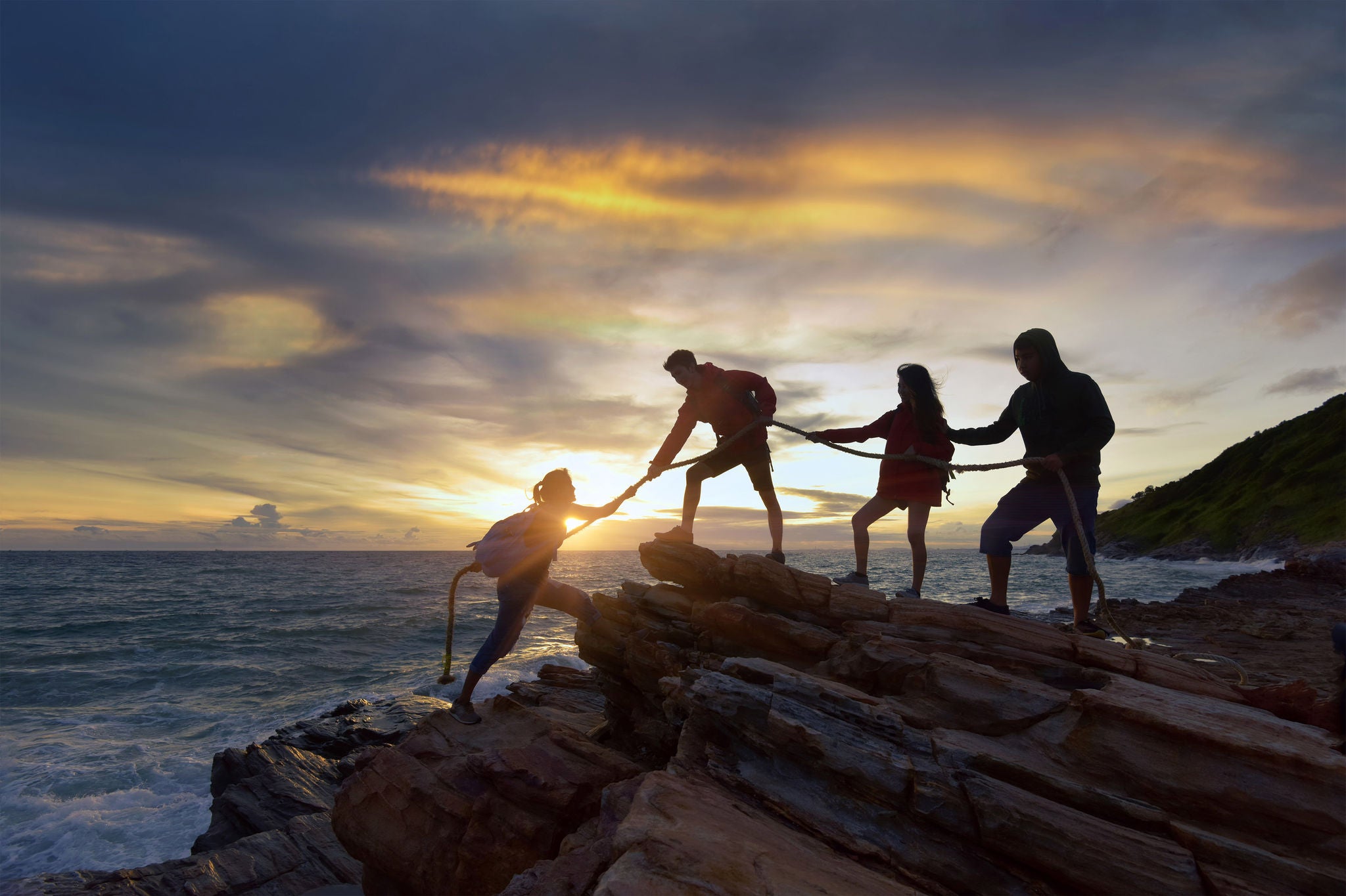 Male and female hikers climbing up silhouette mountain cliff and one of them giving helping hand. People helping and, team work concept.