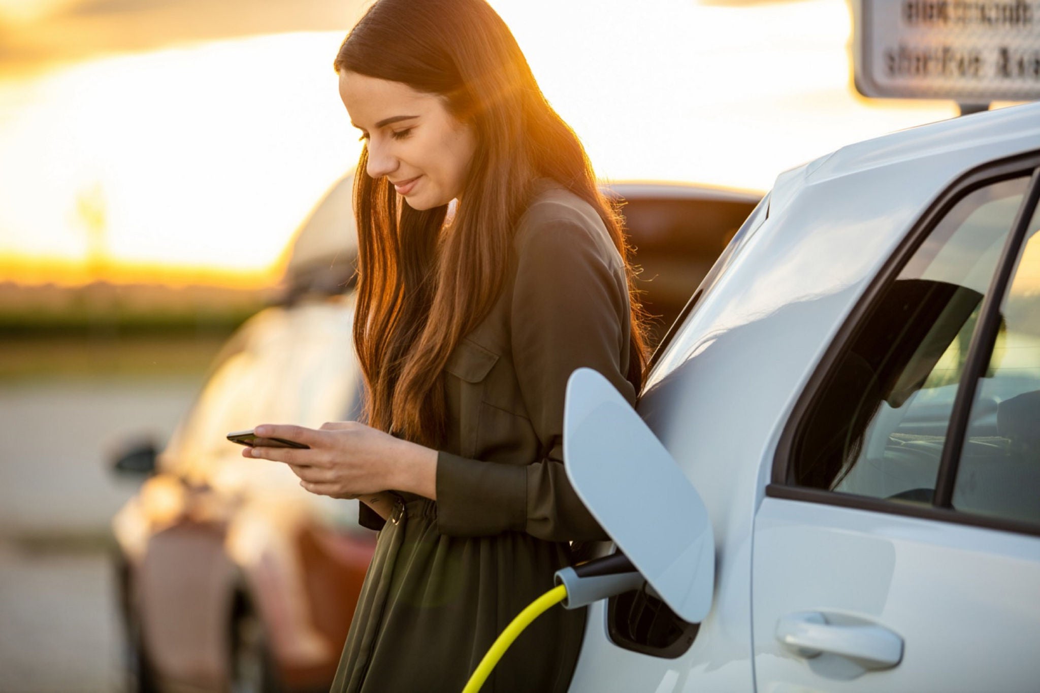 En smilende dame som ser ned på mobilen mens hun lader el-bilen sin.