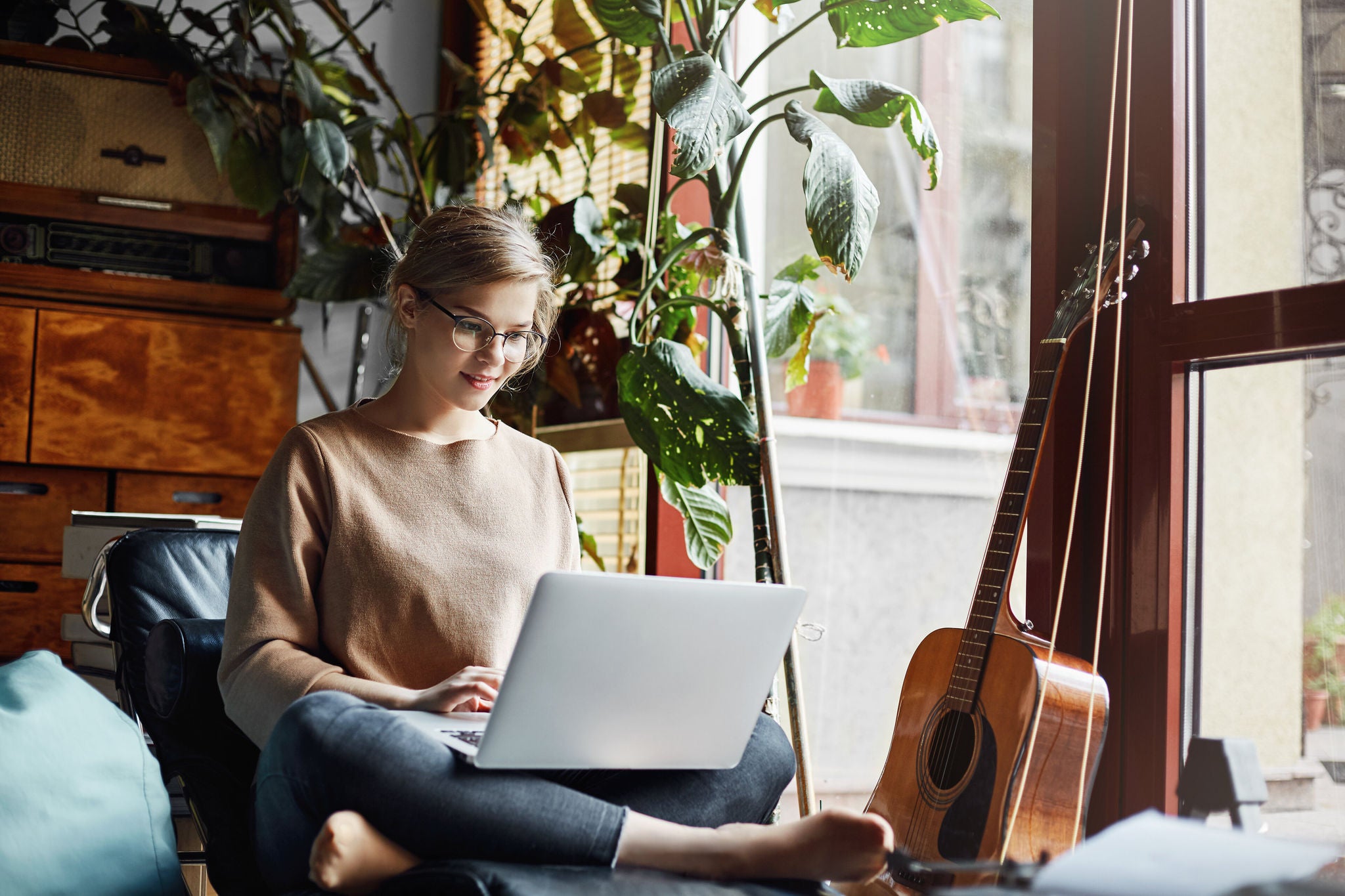 Girl sitting in cozy room near guitar and plants on armchair with crossed feet, holding laptop on laps and typing message, smiling broadly at camera, enjoying working under new design for customer.