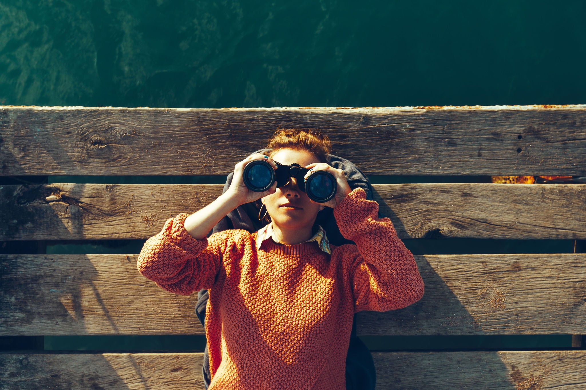 Young Girl Lies On A Pier Near The Sea And Looks Through Binoculars. Travel Search Journey Concept