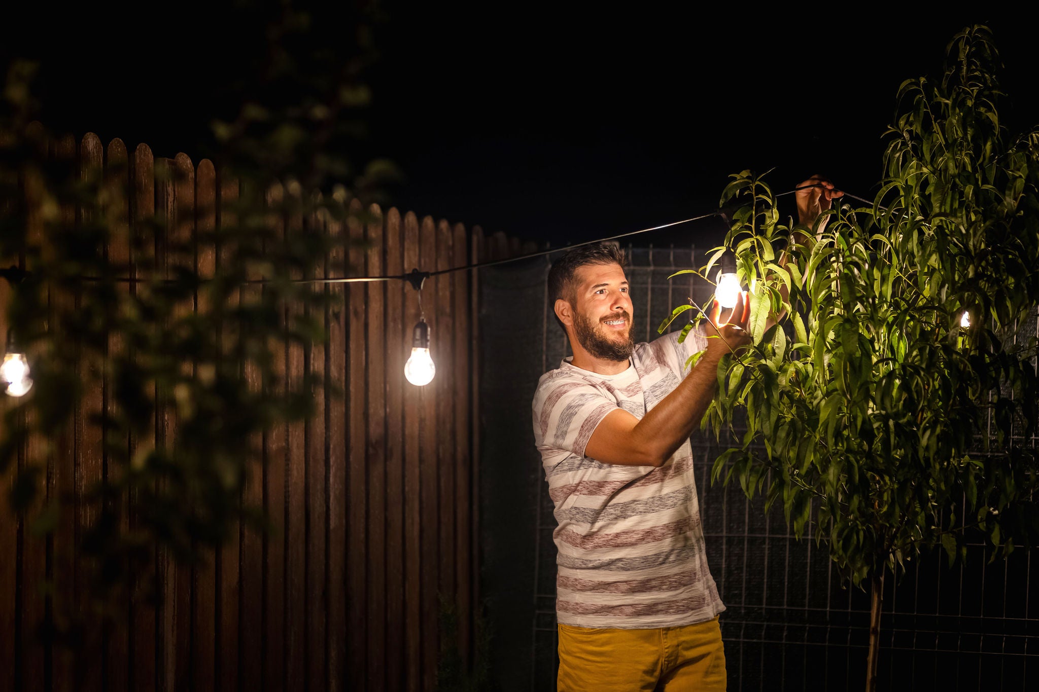 Weekend night mood with smiling millennial guy arranging the light garland for outdoor dinner party in garden