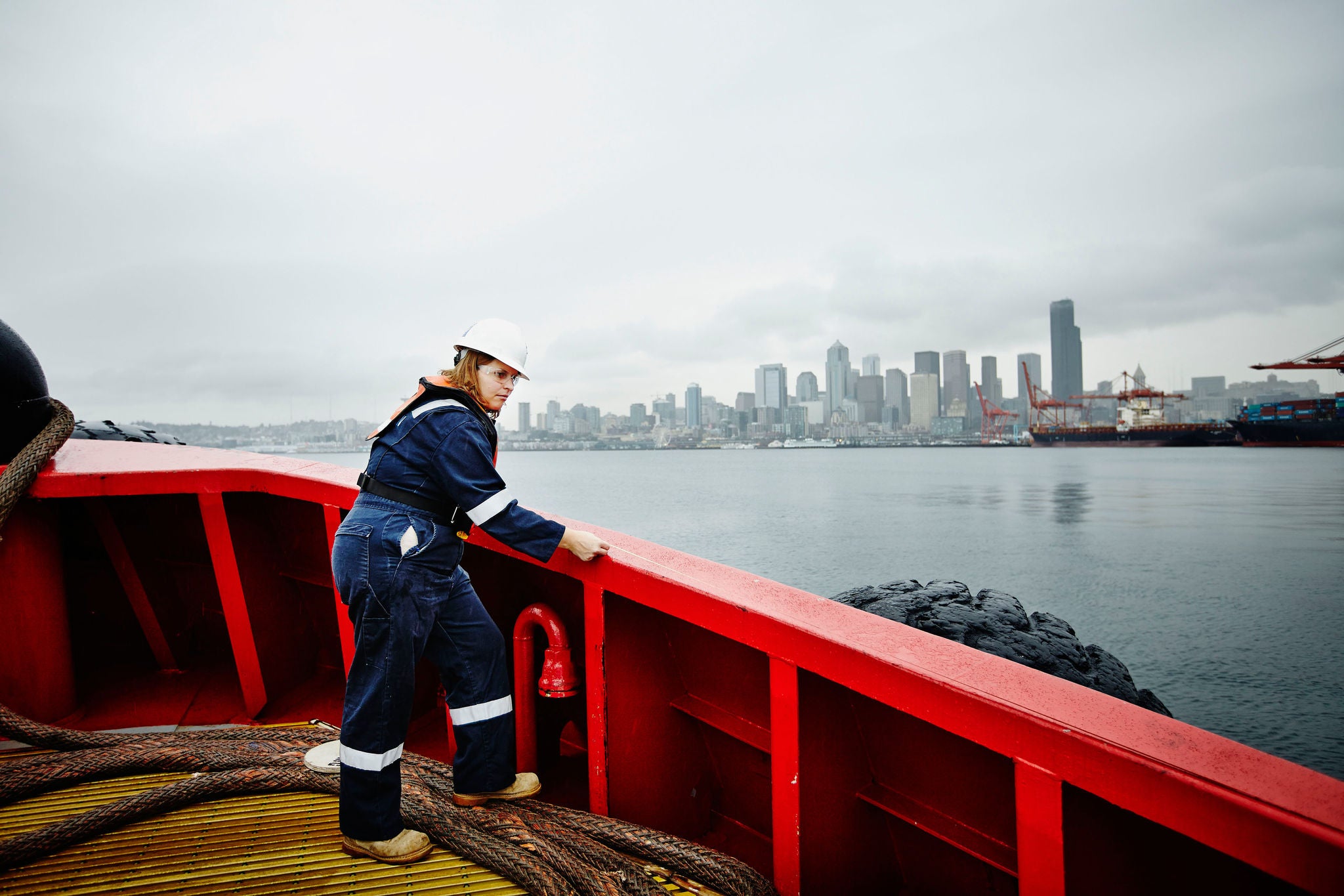 Female naval architect in hardhat and coveralls taking measurement along bulwark of commercial tugboat