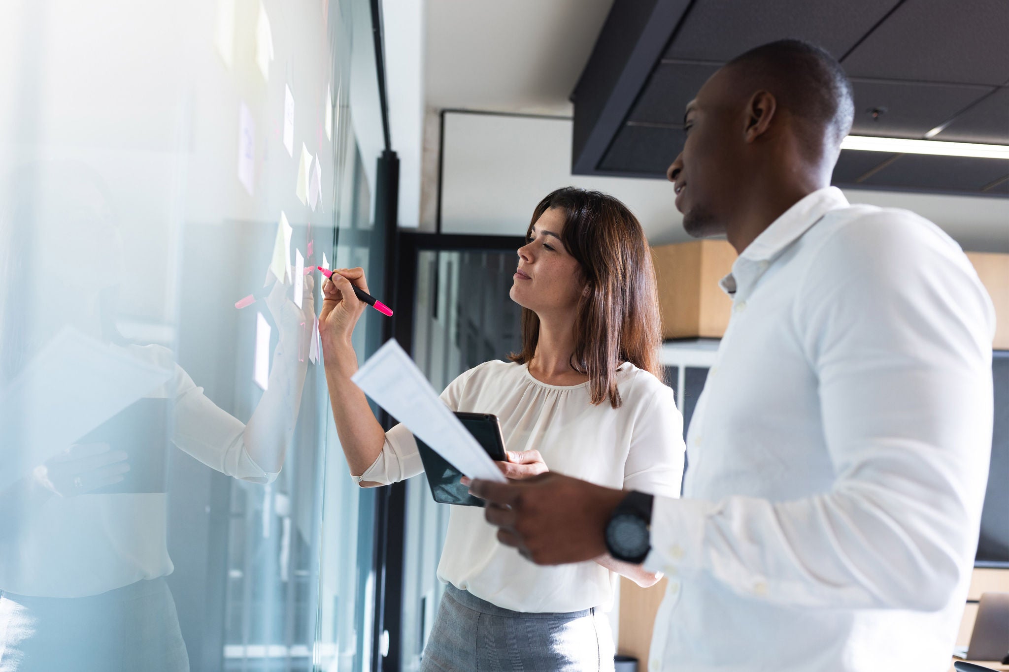 Diverse businessman and businesswoman writing on memo notes on glass board at modern office