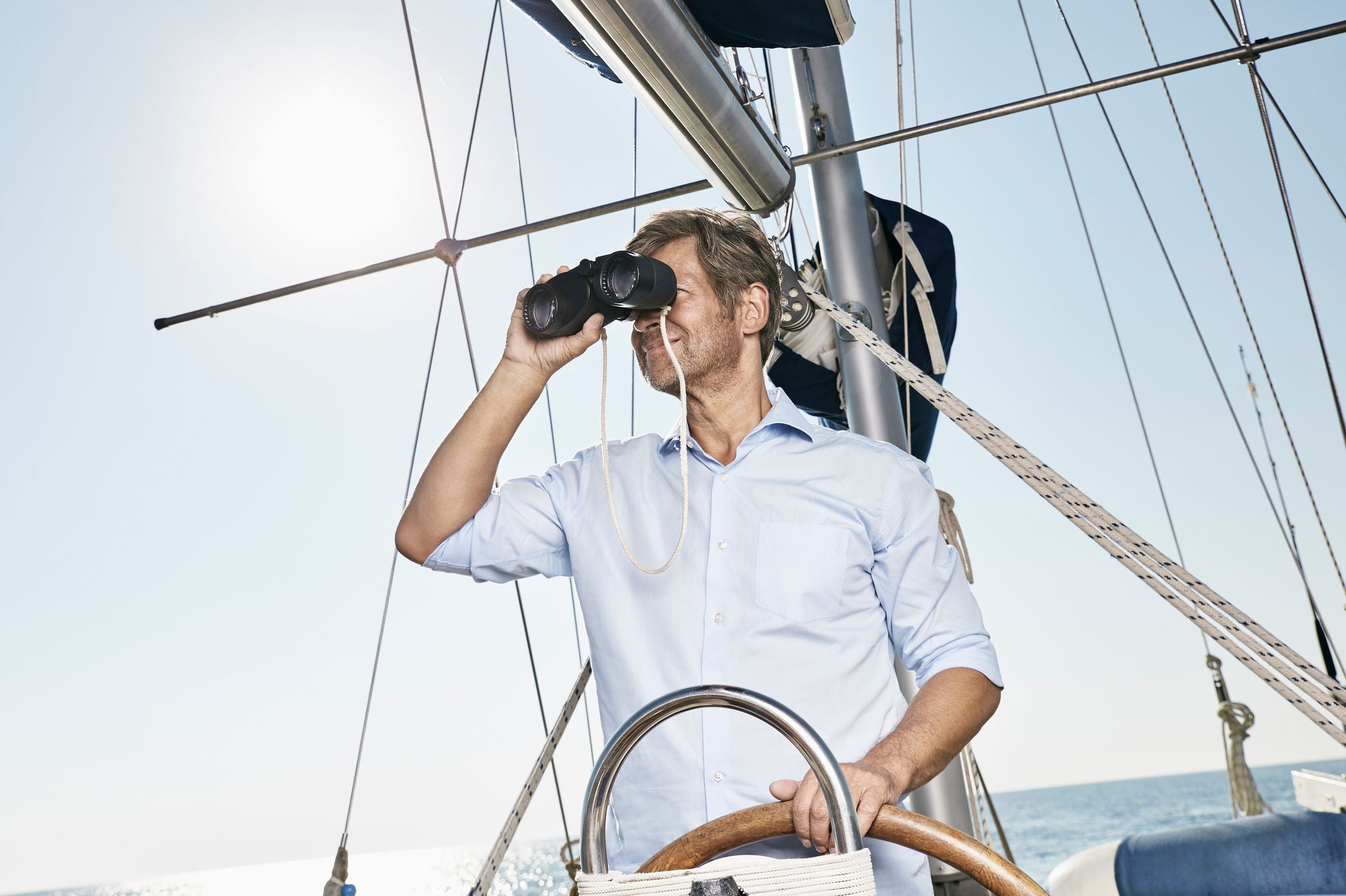 Mature man using binoculars at helm of his sailing boat 