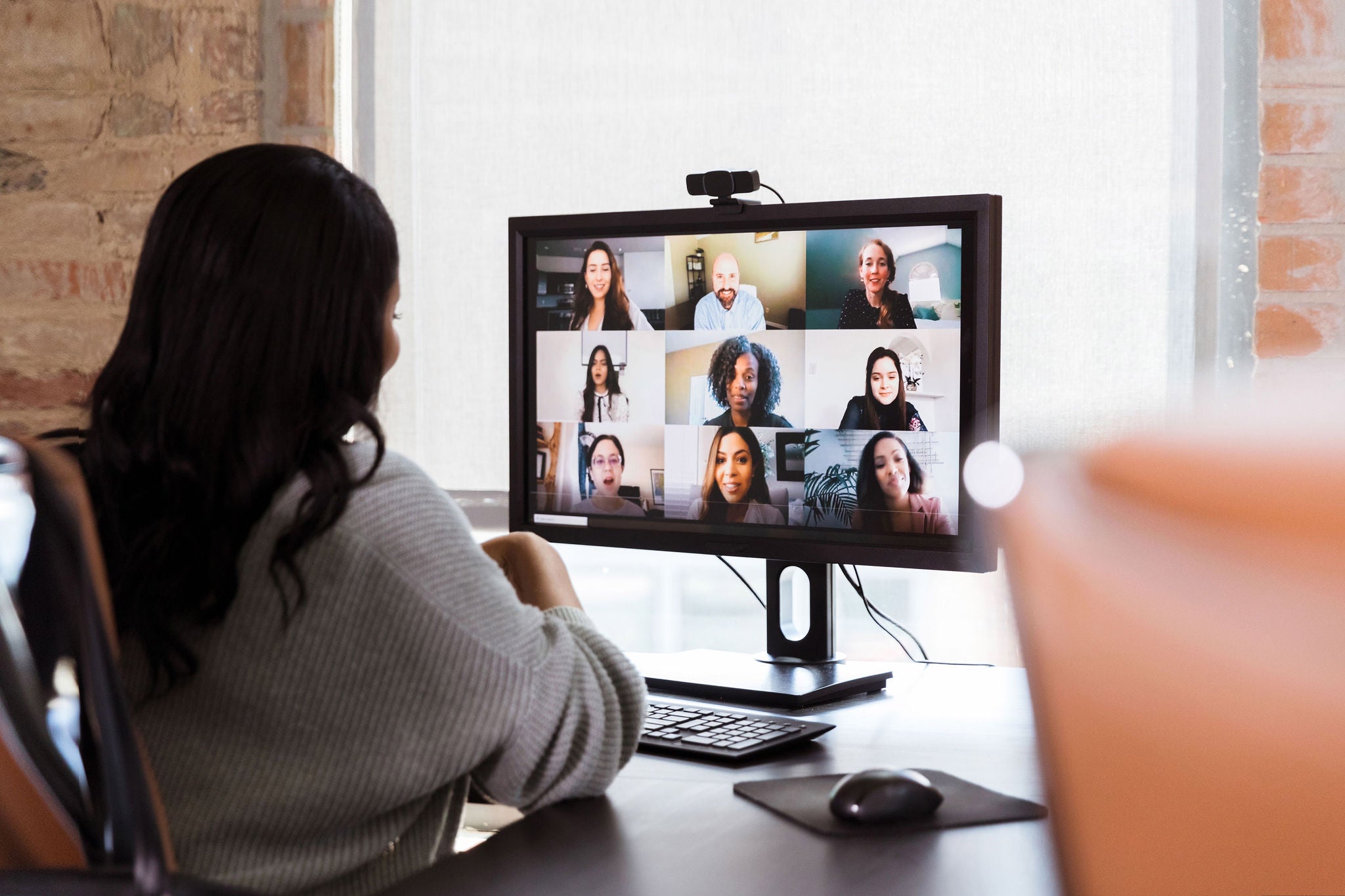 A group of diverse business colleagues participate in a virtual staff meeting during the COVID-19 pandemic. An African American businesswoman participates in the virtual event from her office.