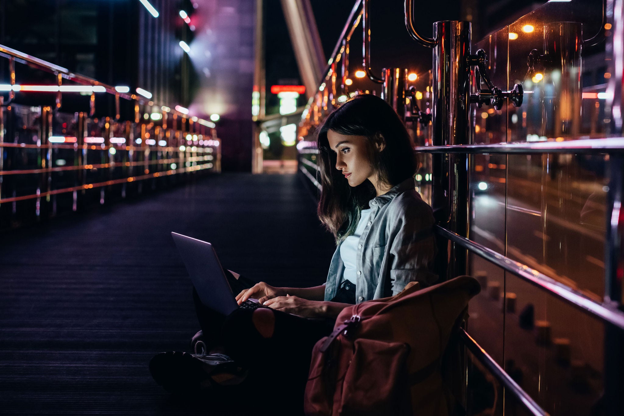 side view of young woman using laptop on street with night city lights on background