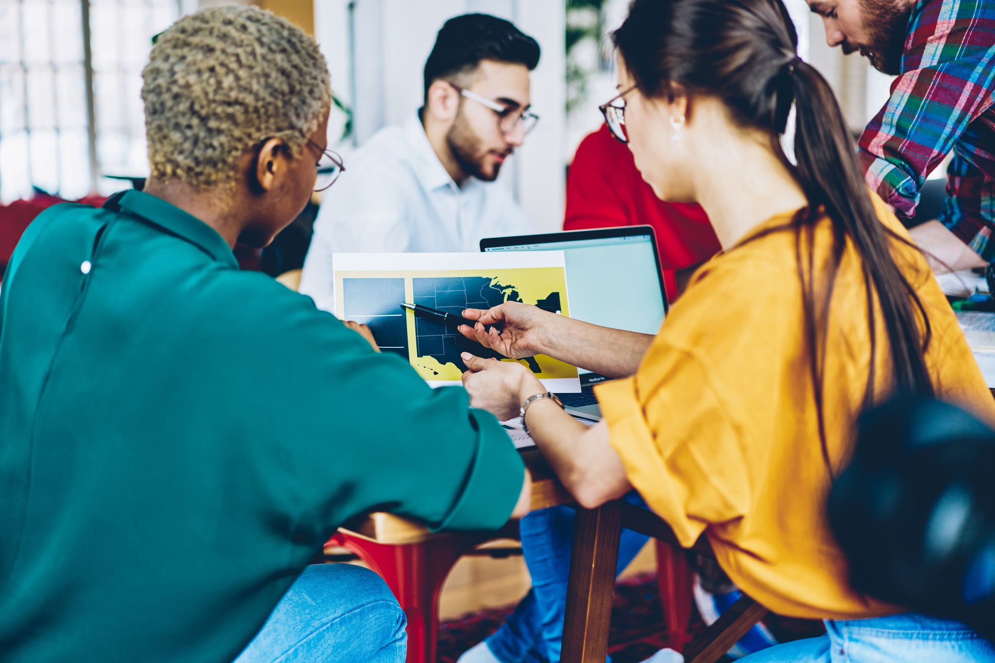 Young professional leader of working team ponting on paper with graphic for improving business plan on meeting table, man coaching multiracial crew of students explaining strategy on workshop