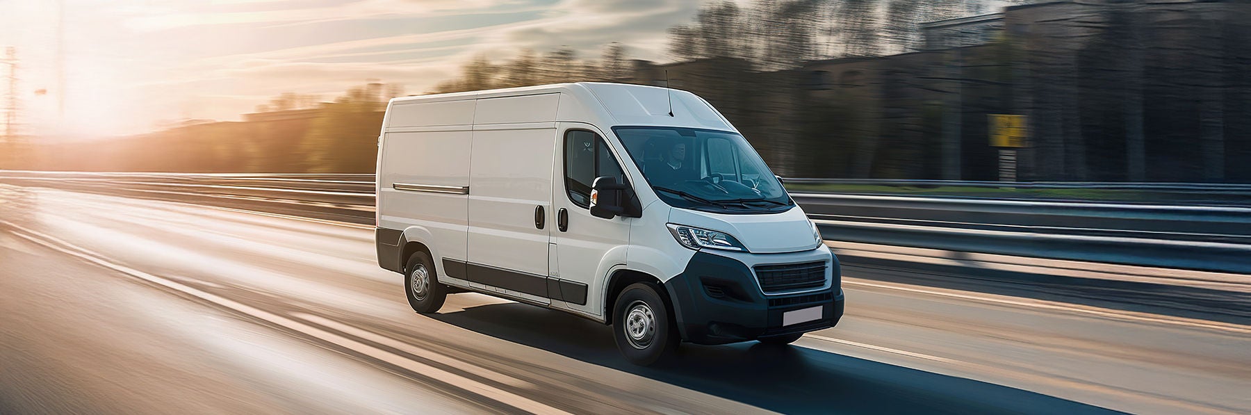 A white van is driving down a road with trees in the background. The van is moving quickly, and the sky is cloudy