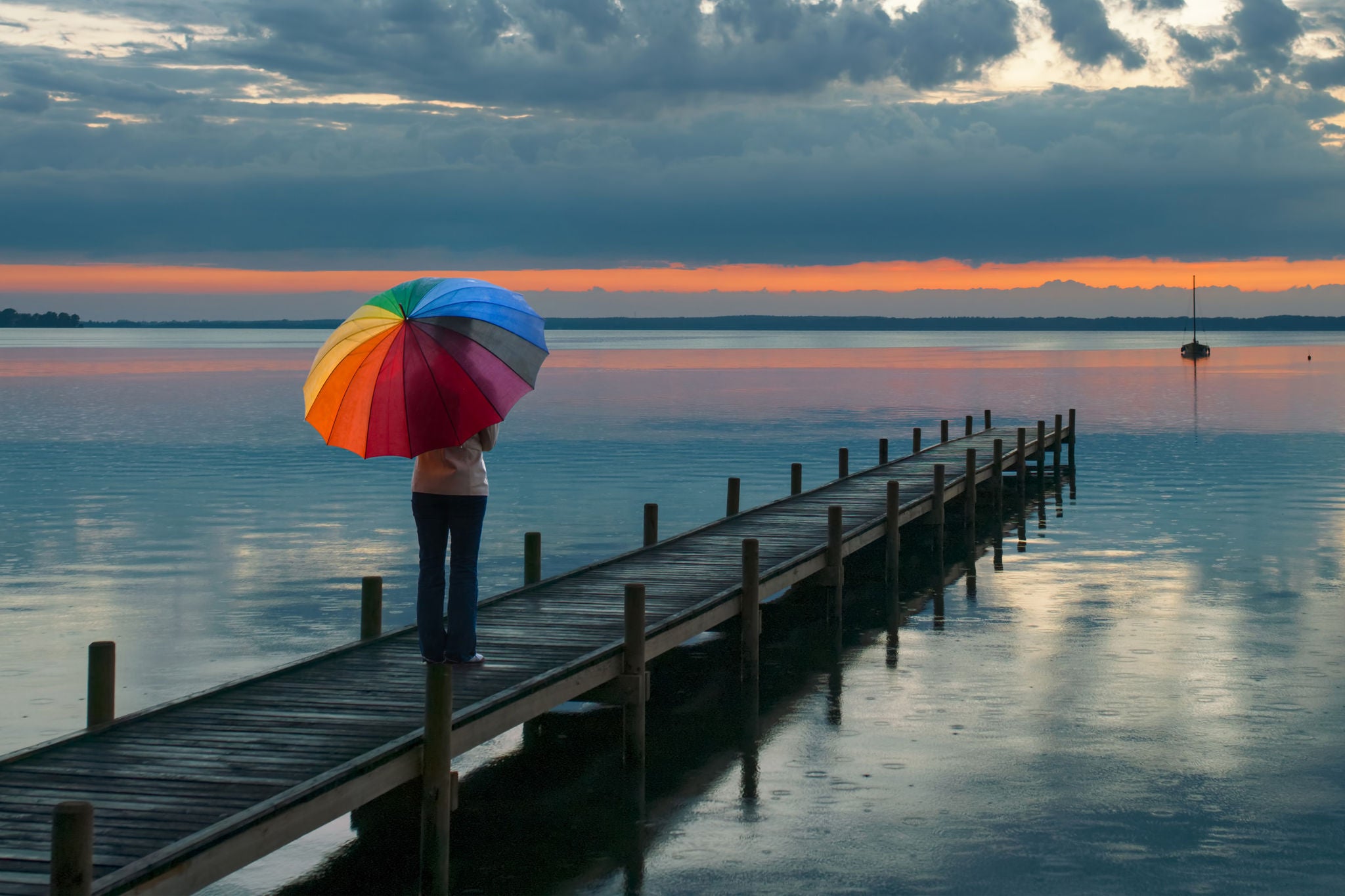 Rear View On Woman With Multi Colored Umbrella In Rain On Lakeside Jetty Watching Reflection Of Majestic Cloudscape At Dusk