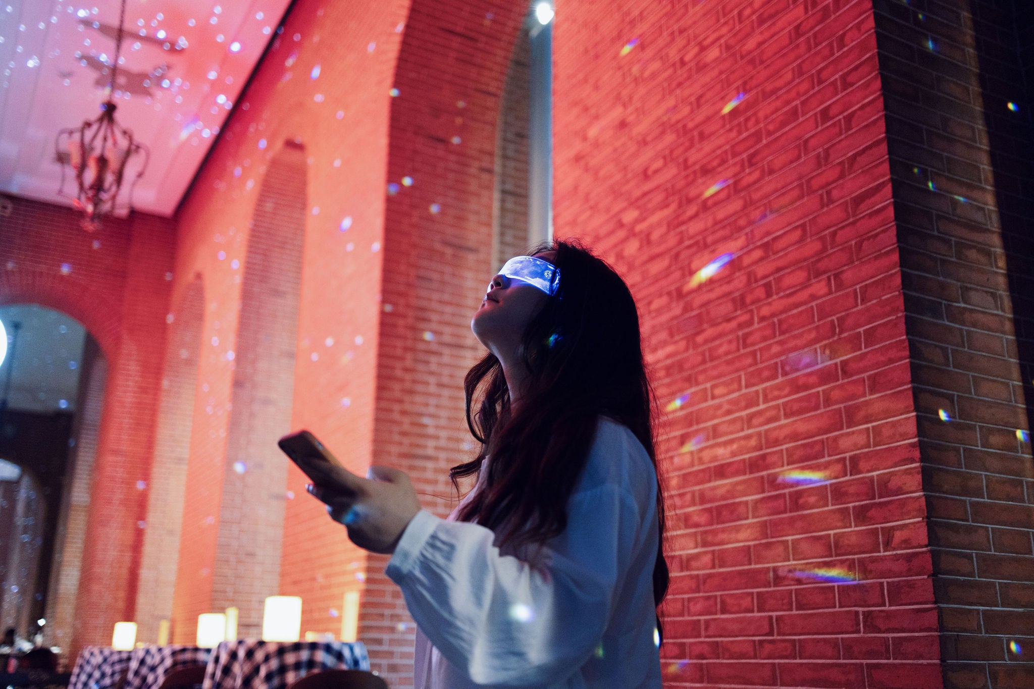 A woman standing in a dimly lit room with brick walls, looking upward while wearing a futuristic, glowing eye mask.