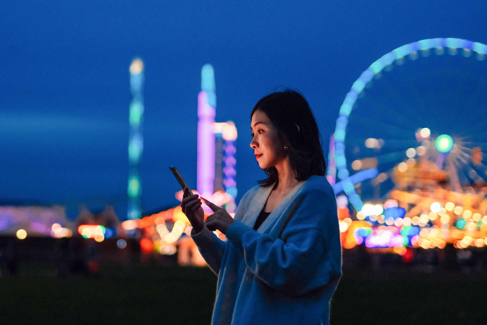 Side view of beautiful young Asian woman looking aways while using smartphone in carnival at night with illuminated night in the background. Mobile banking. Business on the go.