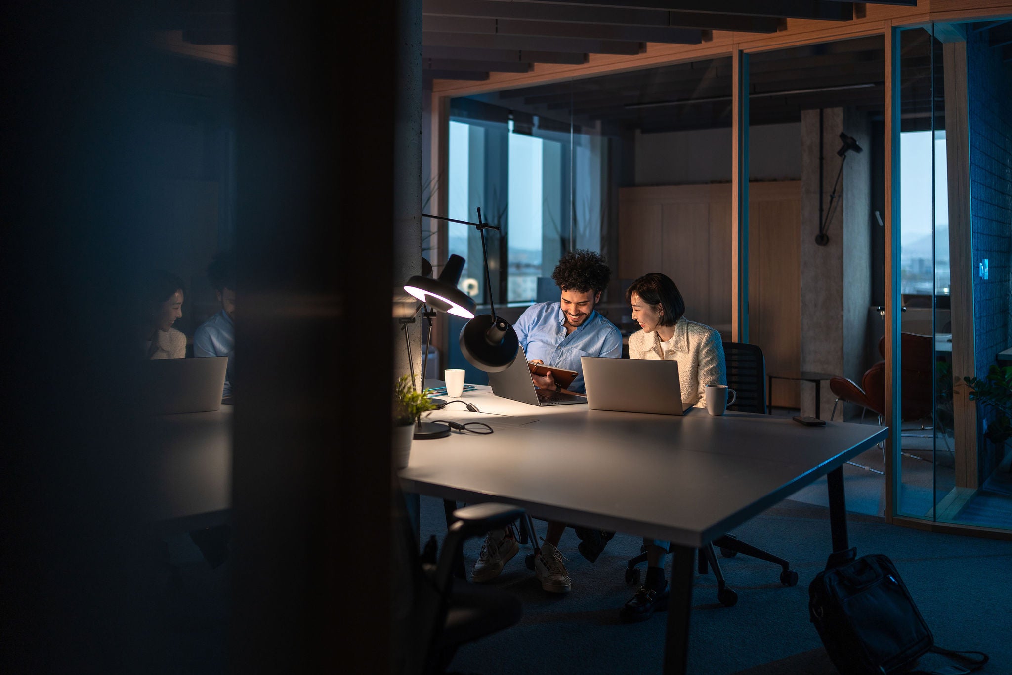 A Middle Eastern businessman and an Asian businesswoman collaborate on a project, focused intently on a laptop in a modern office setting late at night.