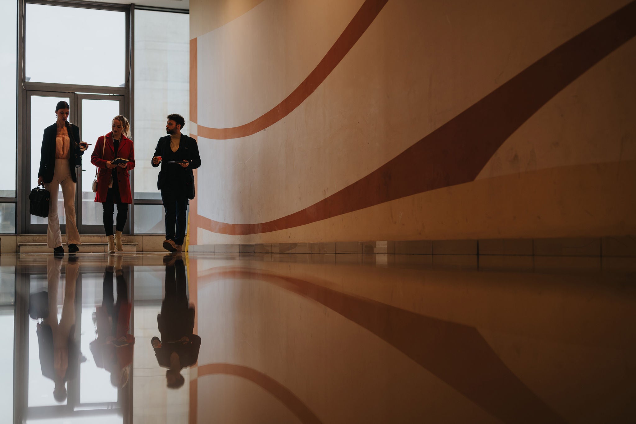 Three business colleagues discussing work while walking in a spacious office corridor with reflective floor and geometric wall pattern.
