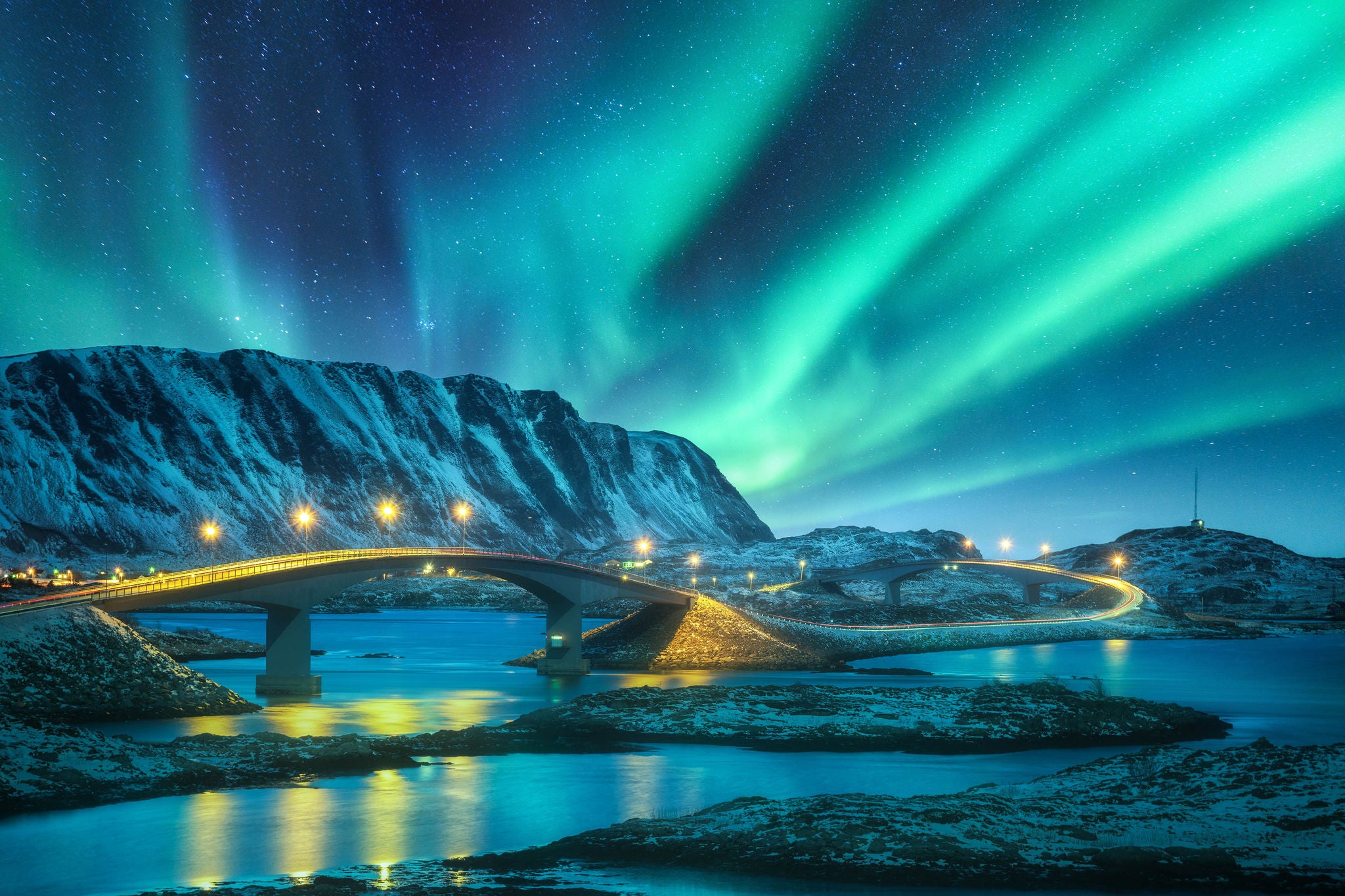 Bridge and northern lights over snowy mountains. Lofoten islands, Norway. Aurora borealis and reflection in water. Winter landscape with starry sky, polar lights, rocks, road, sea, city illumination