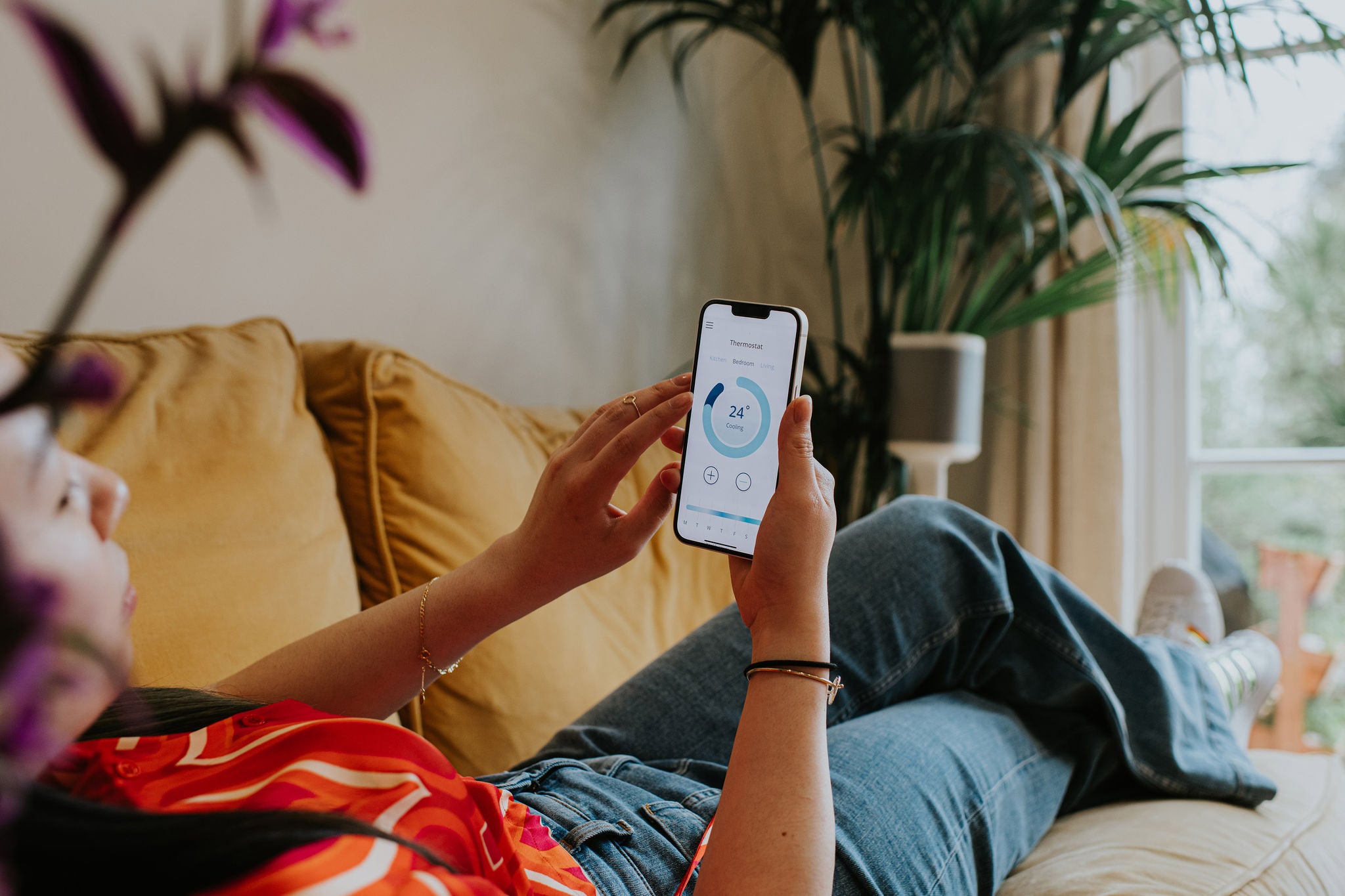 A relaxed woman lies back on a comfortable yellow sofa in a domestic environment. She holds a smart phone and uses a modern thermostat application to control the heating system within the house.