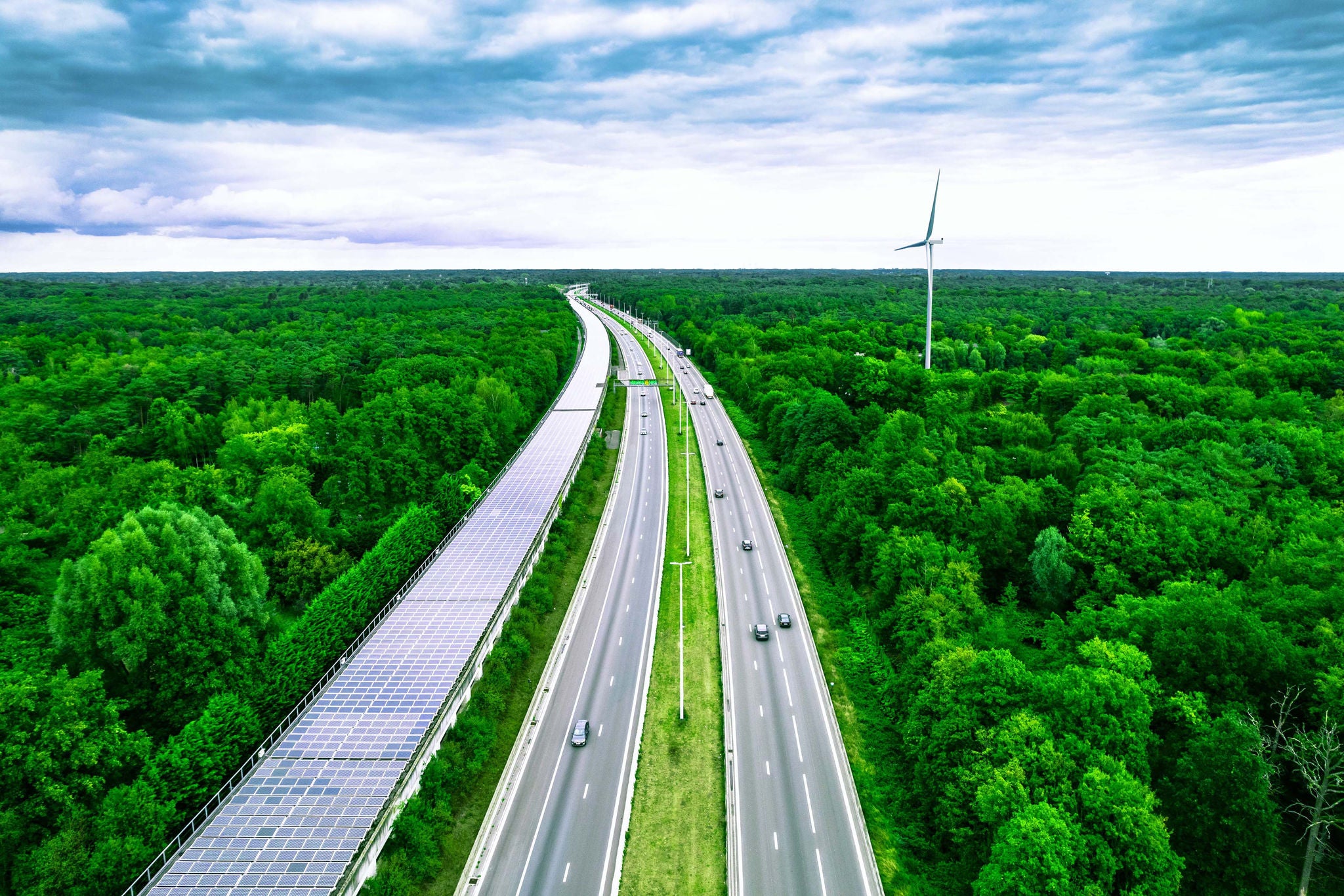 Aerial view taken with drone flying above multiple lane highway crossing luxuriant infinite forest with plain horizon, next to, the Peerdsbos railway tunnel covered with solar pannels with kilometers long. Antwerp. Belgium