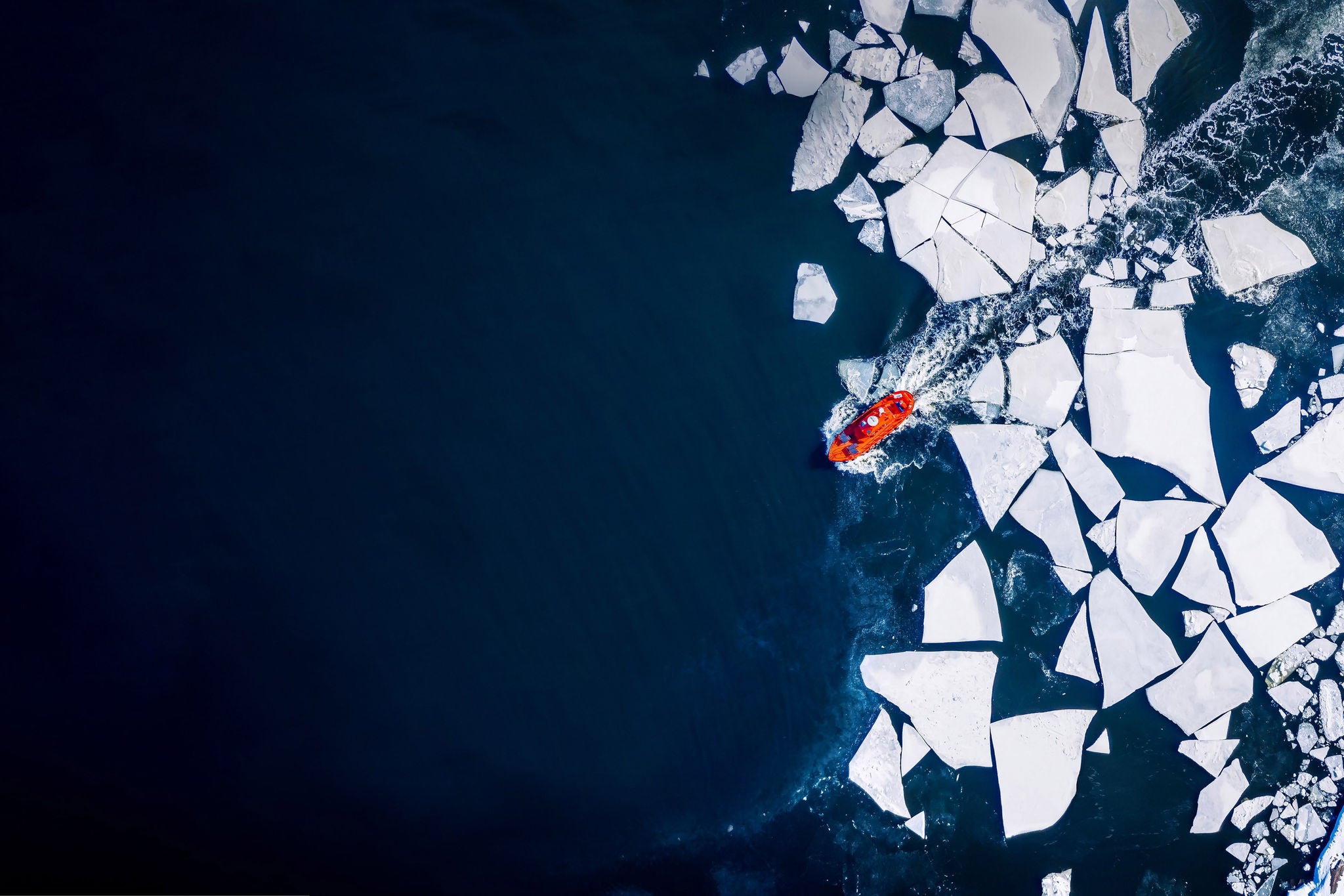 Small orange boat navigating through a dark blue ocean surrounded by large white chunks of ice.