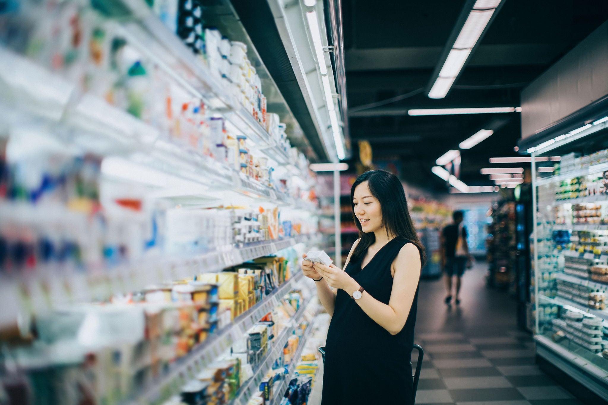 Asian pregnant woman grocery shopping in supermarket and reading nutrition label on a packet of cheese