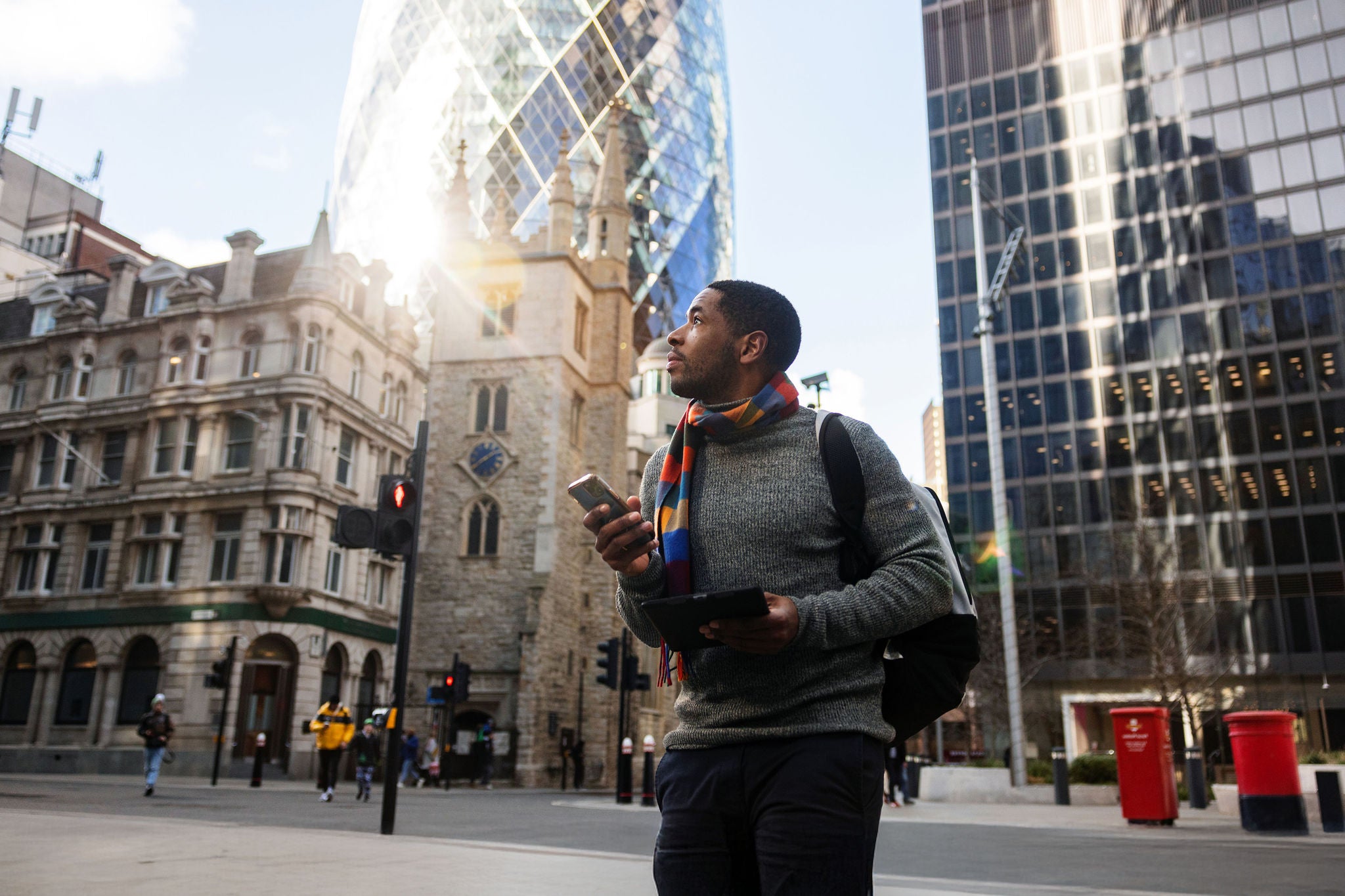 A serious adult black male on his way to a work meeting in London. He is admiring the beautiful streets of London while walking. In his hands, he is carrying a smartphone and a tablet. He looks serious. The weather is nice and sunny. He looks focused.