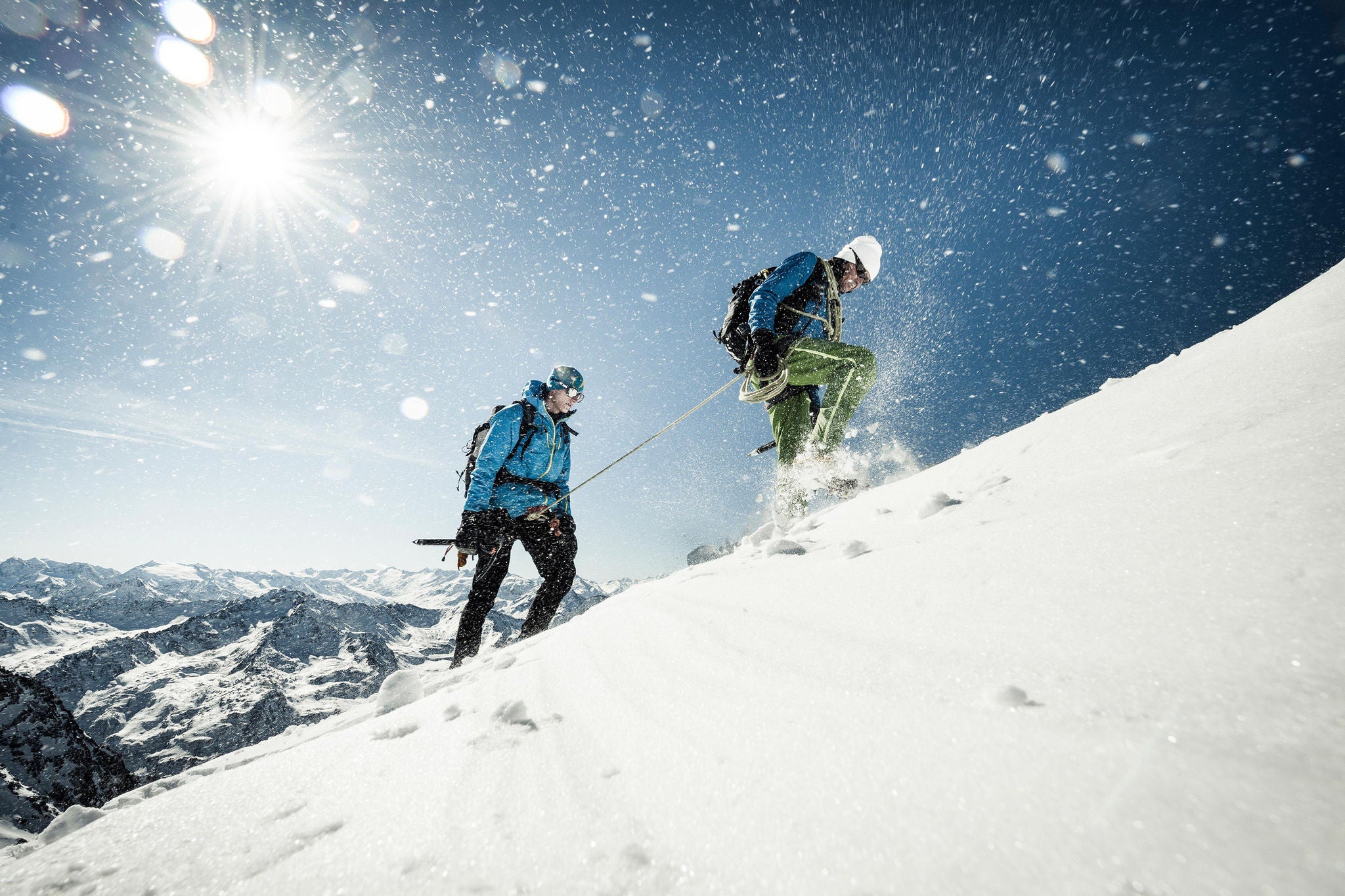 Two climbers hiking up a snowy ridge in strong wind. High mountains in the background.