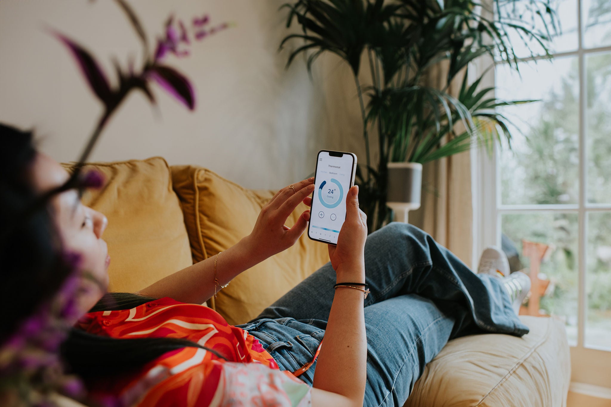 A relaxed woman lies back on a comfortable yellow sofa in a domestic environment. She holds a smart phone and uses a modern thermostat application to control the heating system within the house.