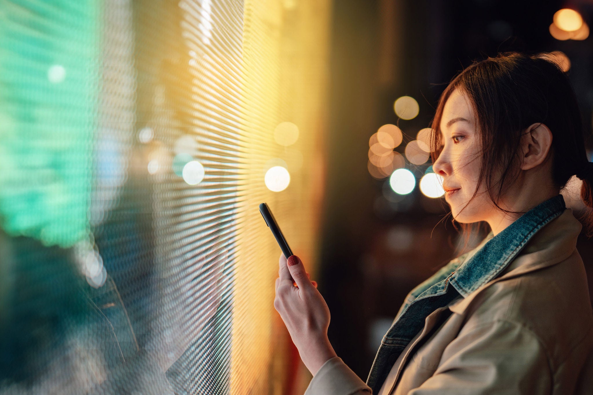 Young Asian woman using smartphone against illuminated digital display in city street at night. Blockchain technology. Trading cryptocurrency. NFT (Non-Fungible Token) investment.