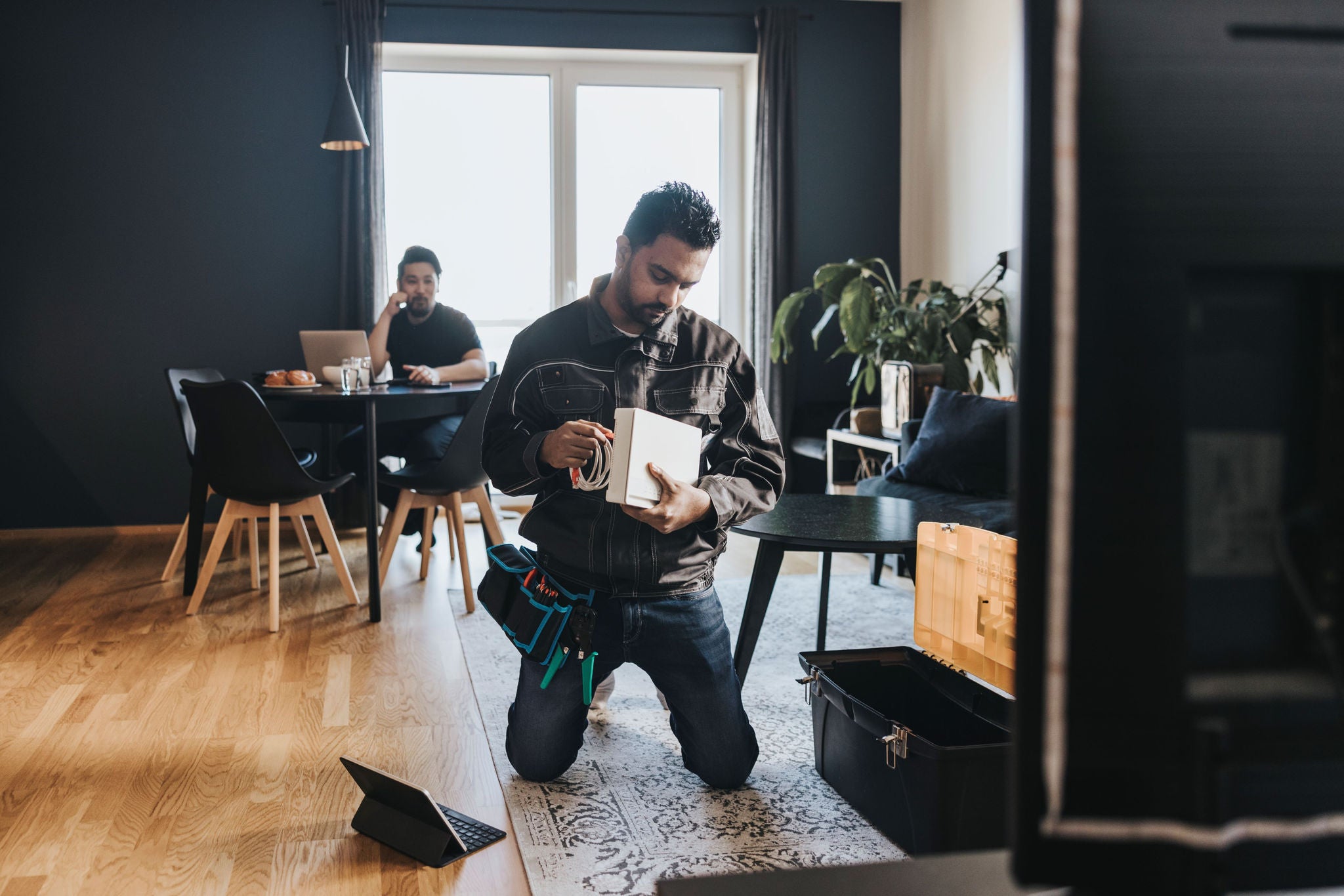 A person kneeling on the floor in a modern living room, holding a device and cables, likely performing technical or repair work. Another person is seated at a dining table, working on a laptop. The room features wooden flooring, a dark accent wall, large windows, indoor plants, and a coffee table with a toolbox.