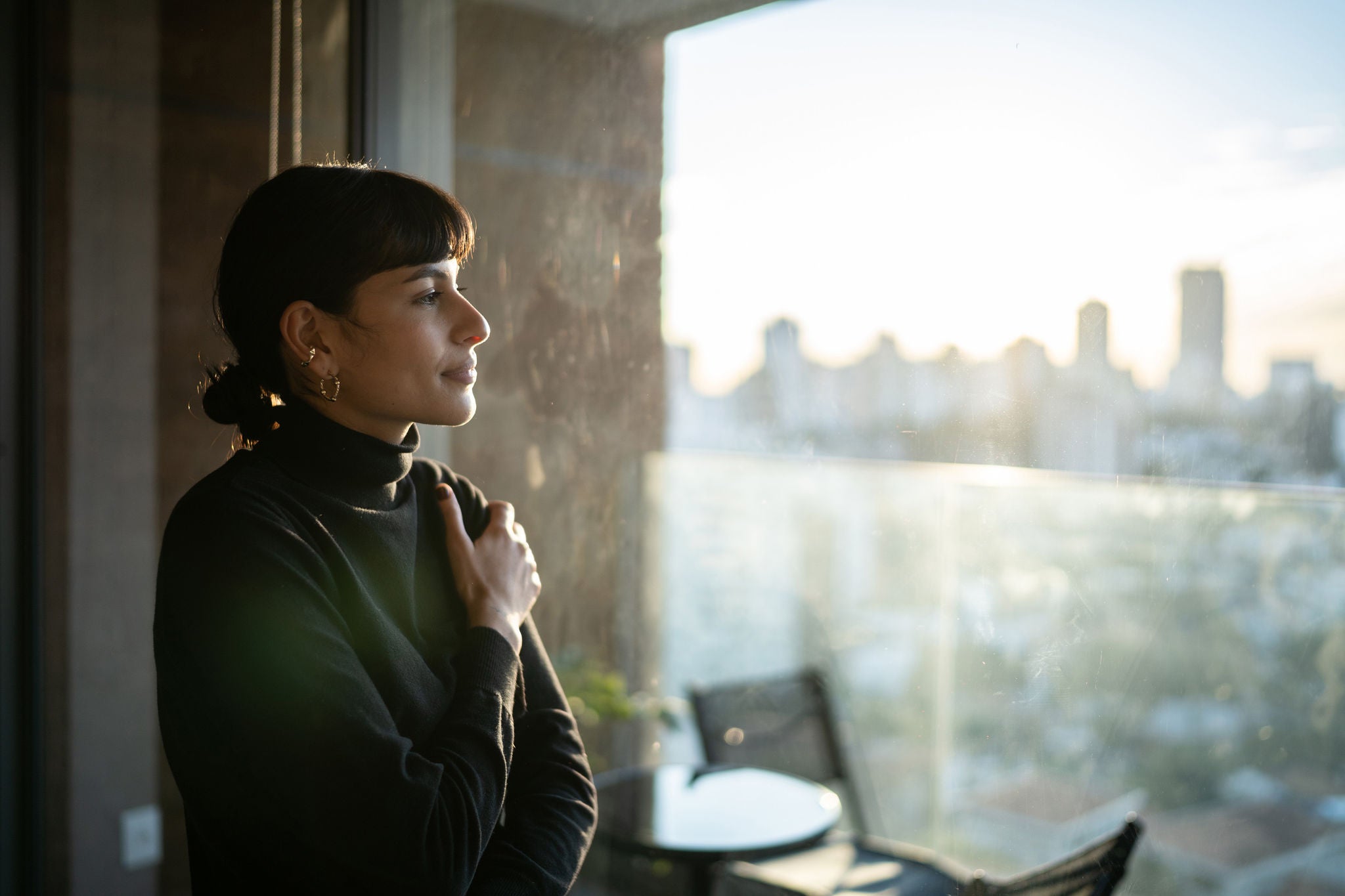 A woman standing near a window in a softly lit room, gazing thoughtfully outside at a city skyline during sunset.