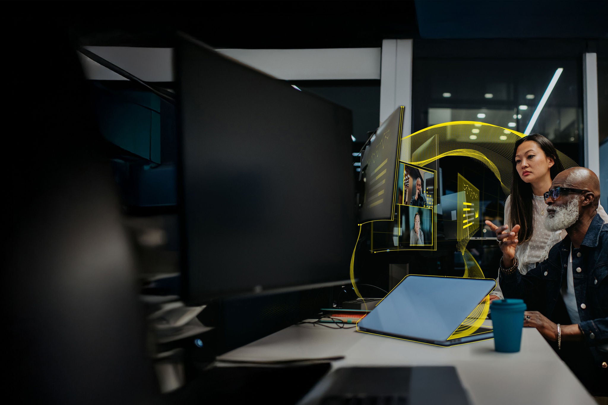 A older male and a young female employee work late in a modern office space. The woman stands behind the man, as they both look a multiple computer screens. It is dark outside suggesting they are working late into the night. They are participants on an online video call.