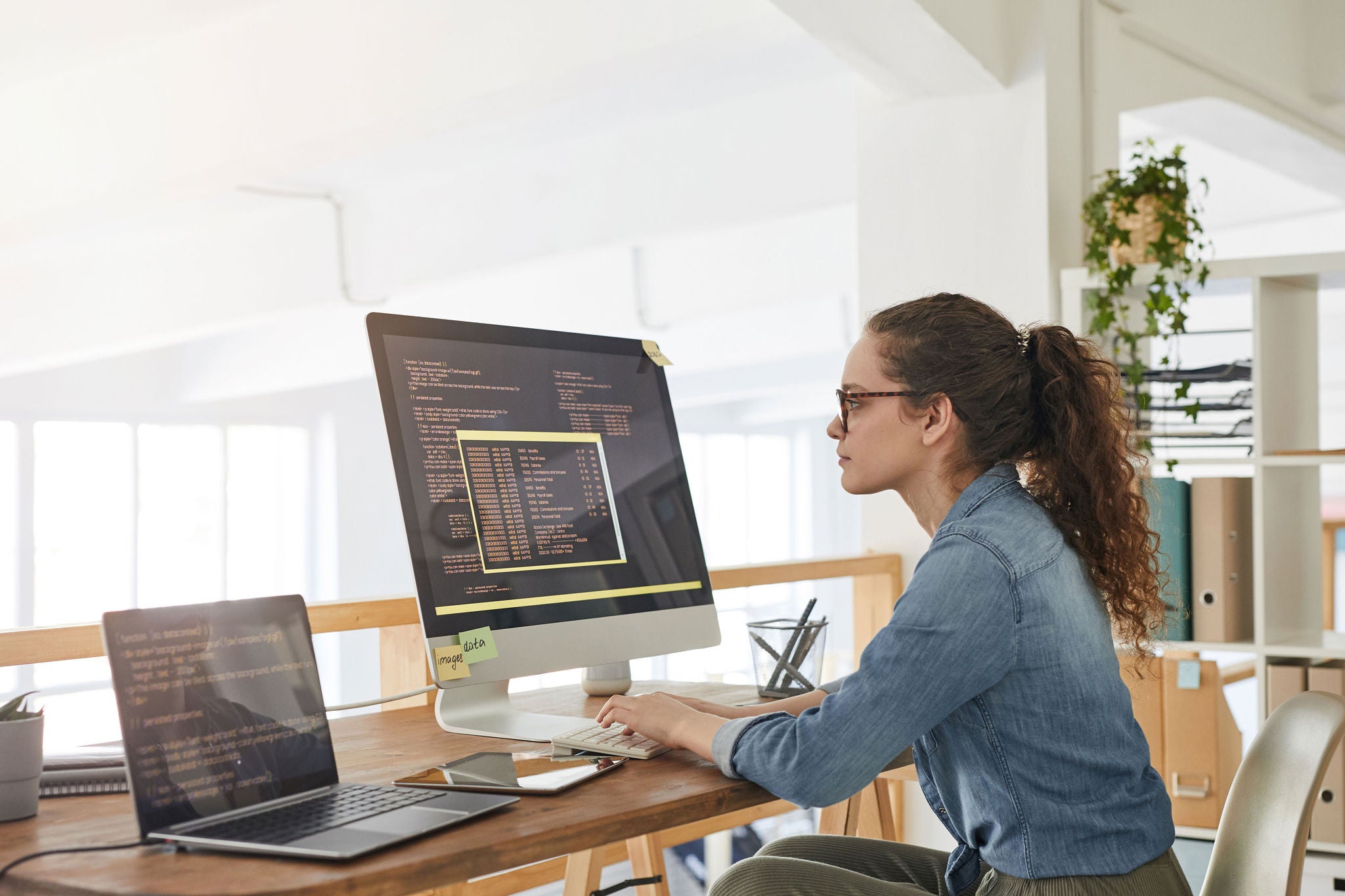 Side view portrait of female IT developer typing on keyboard with black and orange programming code on computer screen and laptop in contemporary office interior, copy space