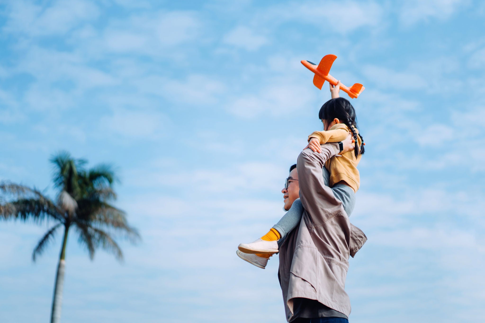 Joyful young Asian father carrying his little daughter on shoulders, spending time together outdoors, playing with airplane toy in park on a lovely sunny day against beautiful blue sky