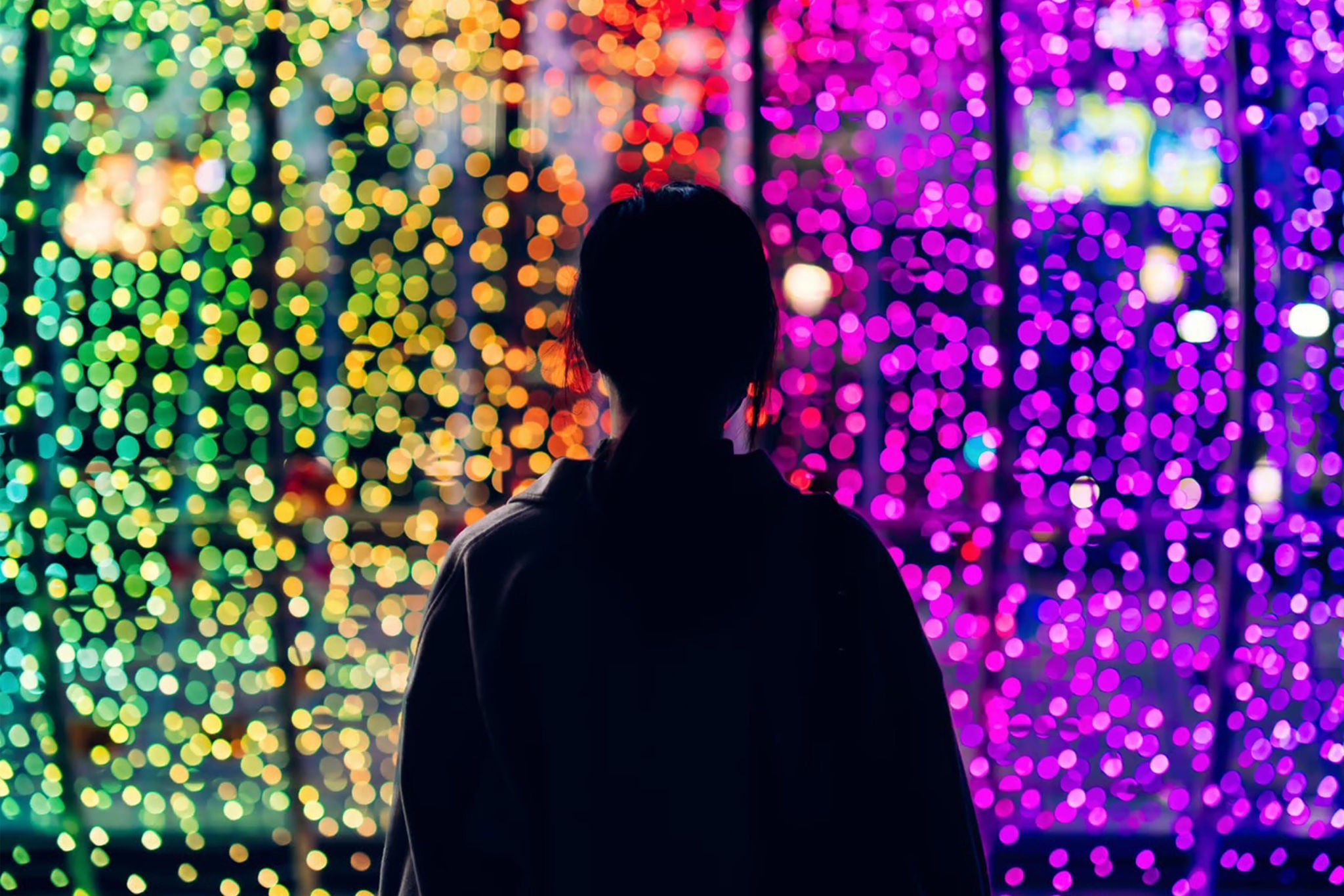 Silhouette of young woman standing against illuminated and colourful bokeh lights background in the city at night - stock photo