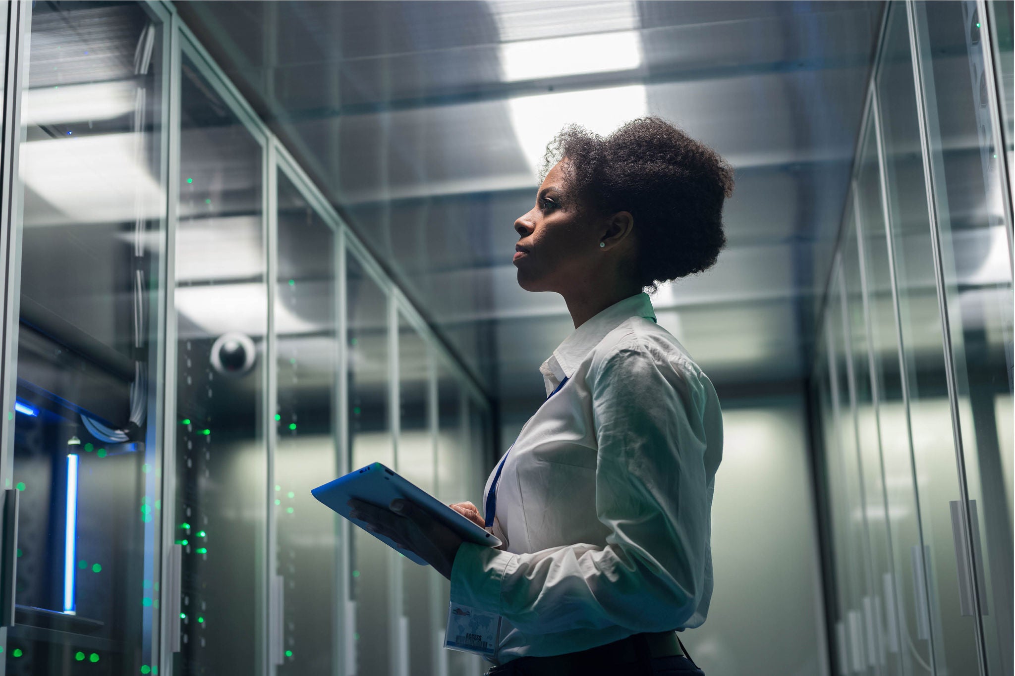 A woman in a white shirt holding a tablet is standing in a server room, looking intently at the servers