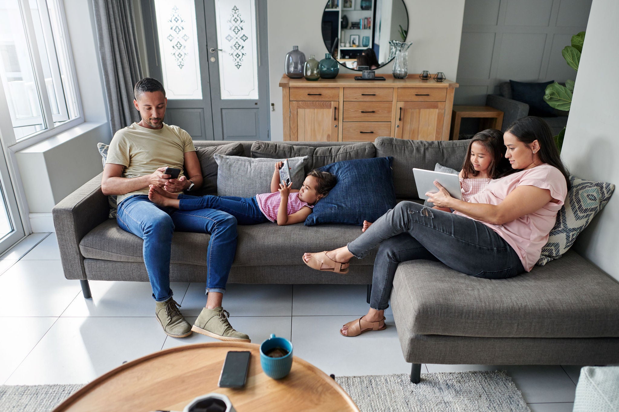 A family of four sitting on a gray sectional sofa in a modern, well-lit living room, using electronic devices. The room has large windows, a wooden sideboard with decorative items, and a round wooden coffee table.