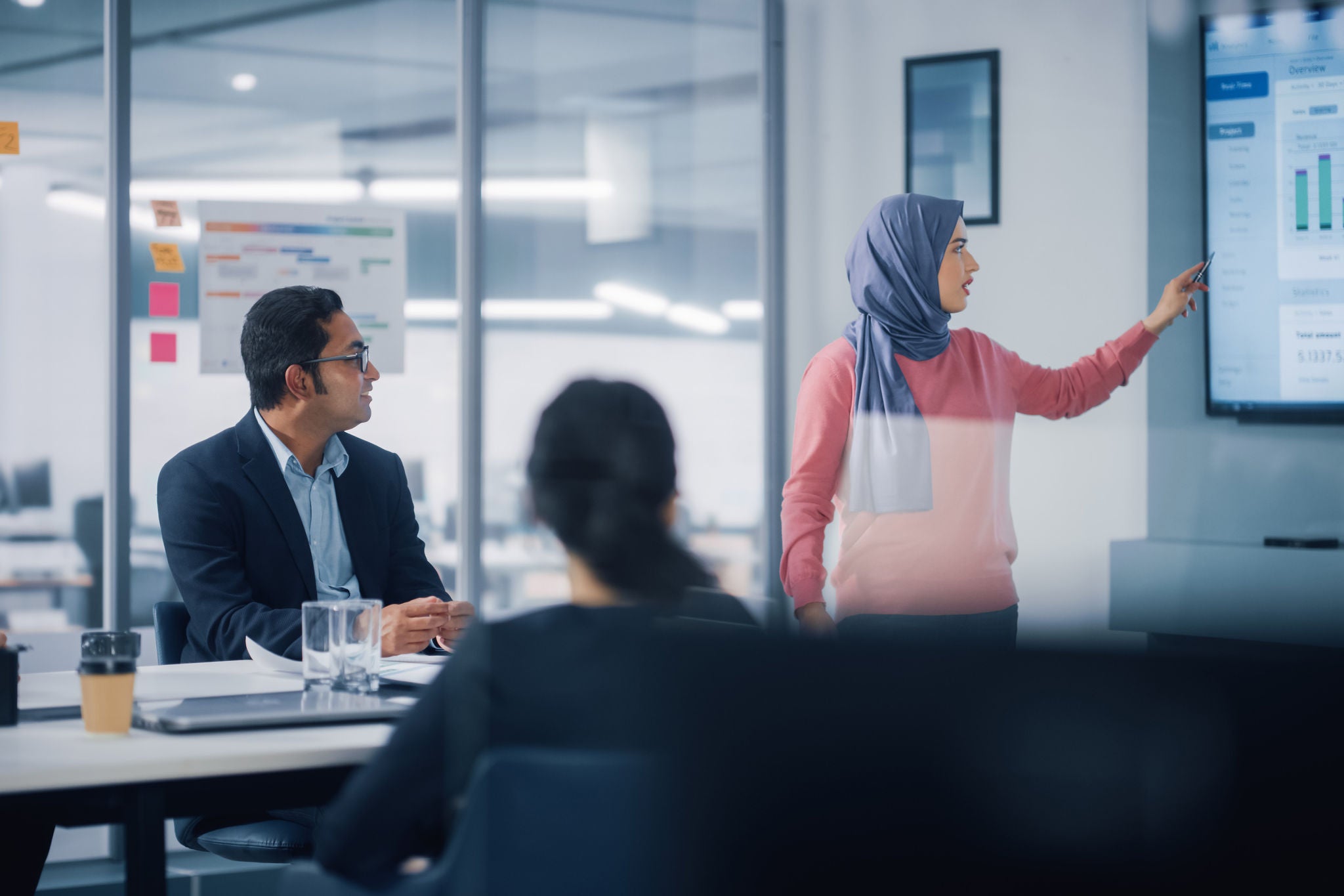 Multi-Ethnic Office Conference Room. Brilliant Muslim Female CEO Wearing Hijab does Presentation for Group of Managers Talking, Using TV Infographics, Statistics. Innovative Businesspeople.