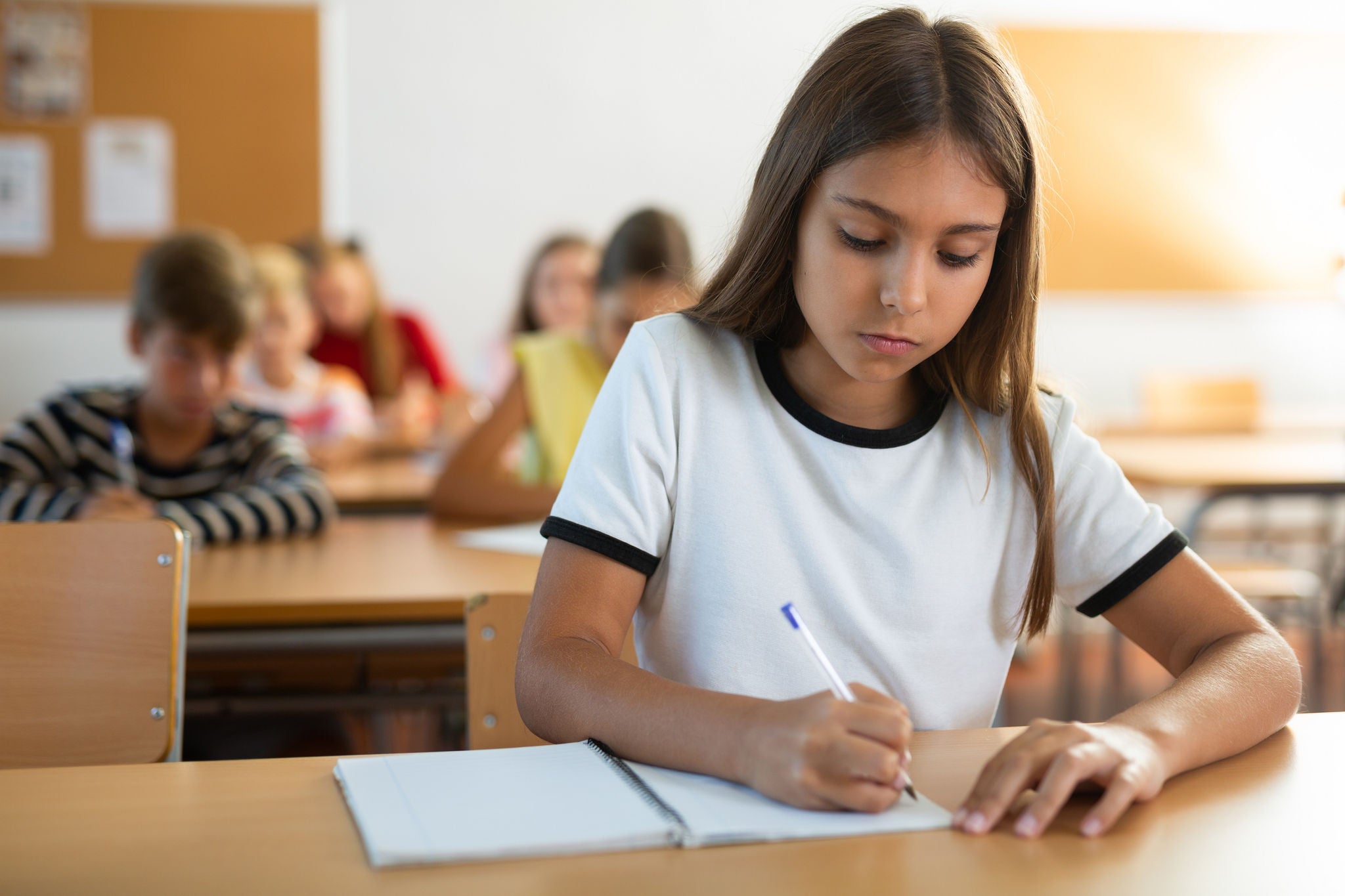 Young girl sitting at desk with her classmates during lesson in school and writing in copybook.