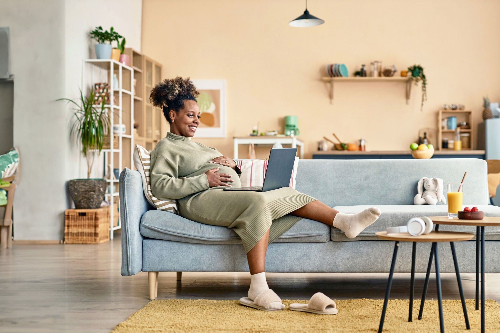 Wide shot of happy pregnant African American woman lying back on big couch in living room with laptop on lap while embracing her round belly