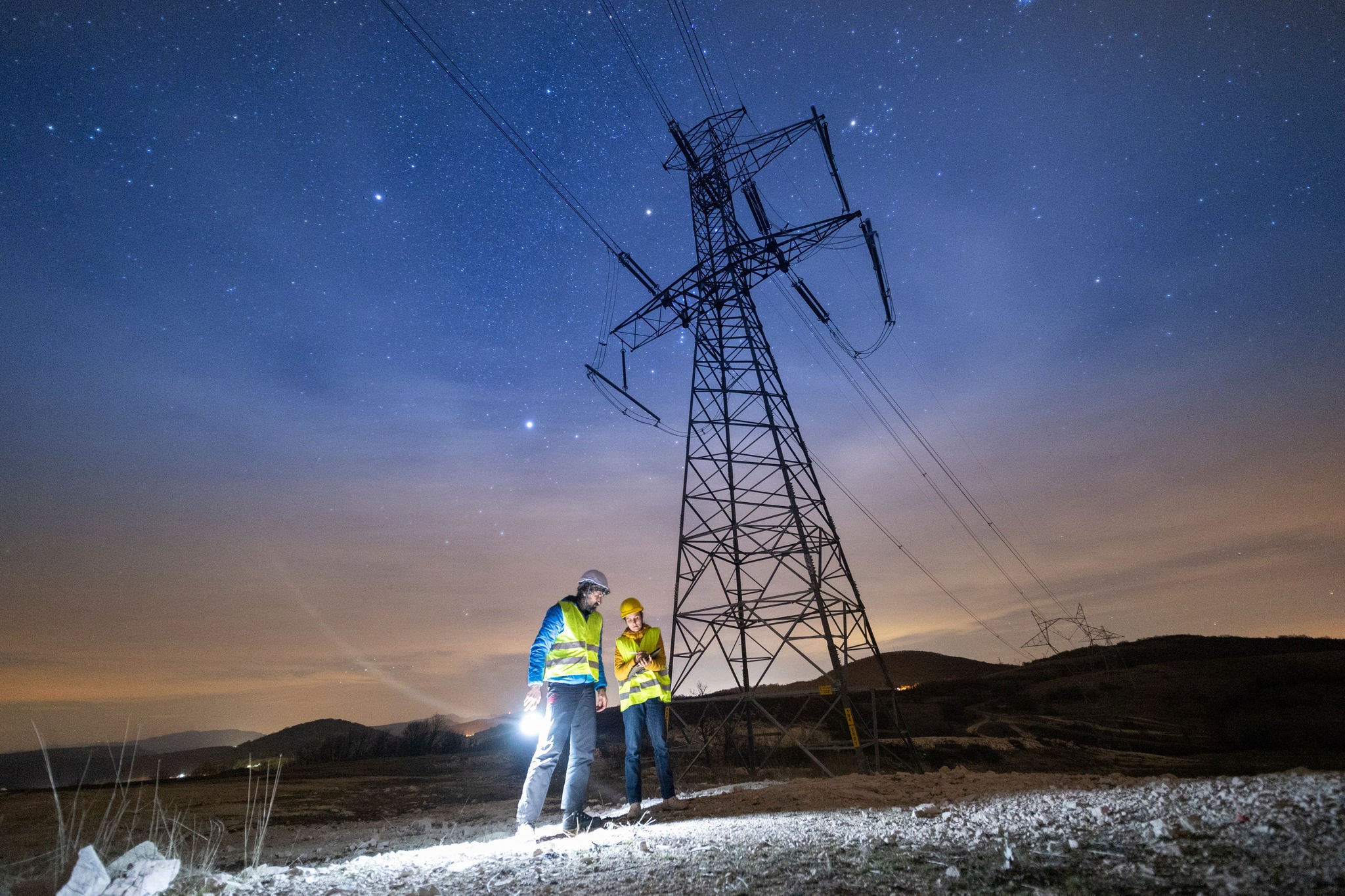 Two workers wearing safety vests and helmets inspect the ground under a large power transmission tower at night