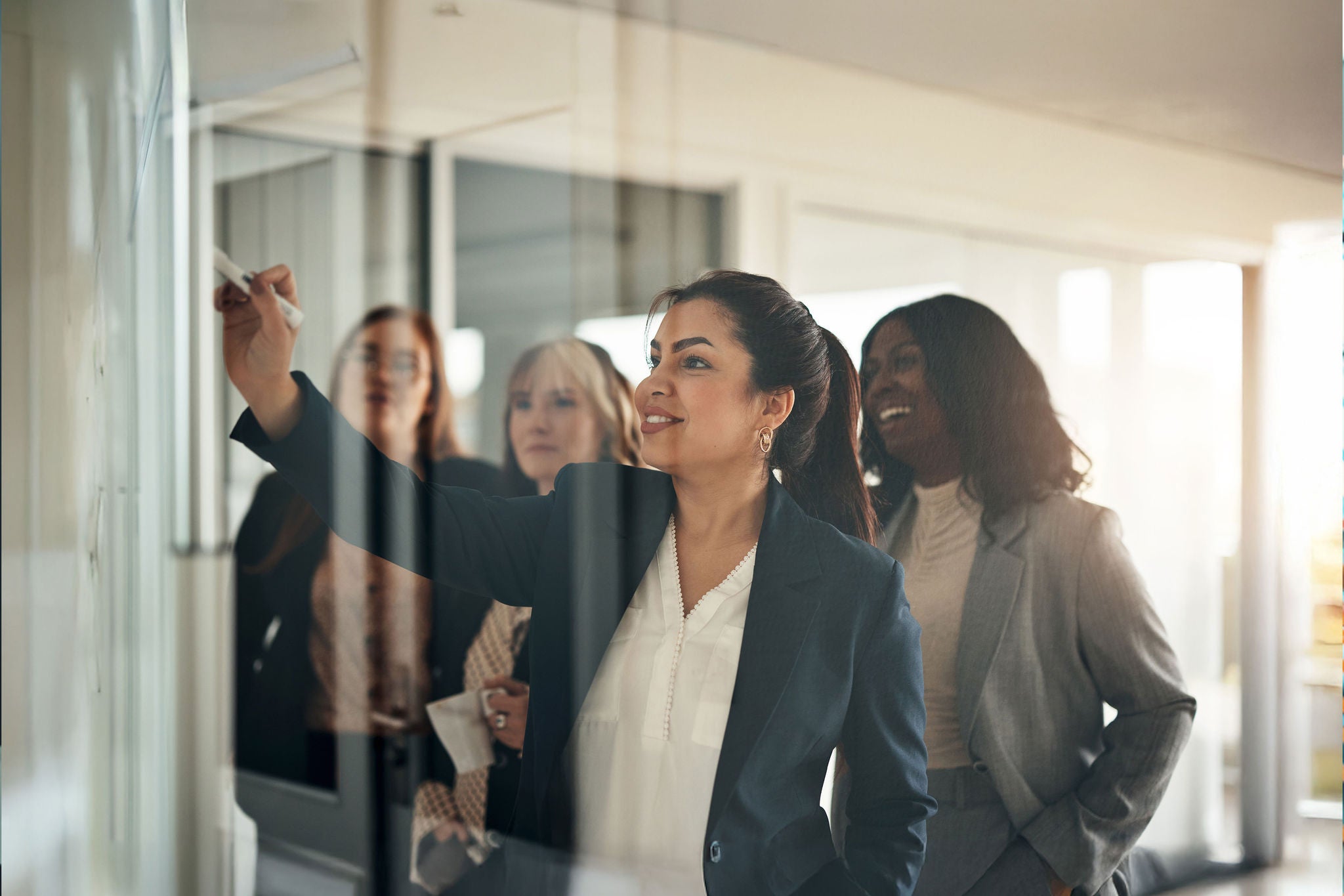 Smiling female executive and a diverse team of businesswomen brainstorming together on a whiteboard inside of an office boardroom