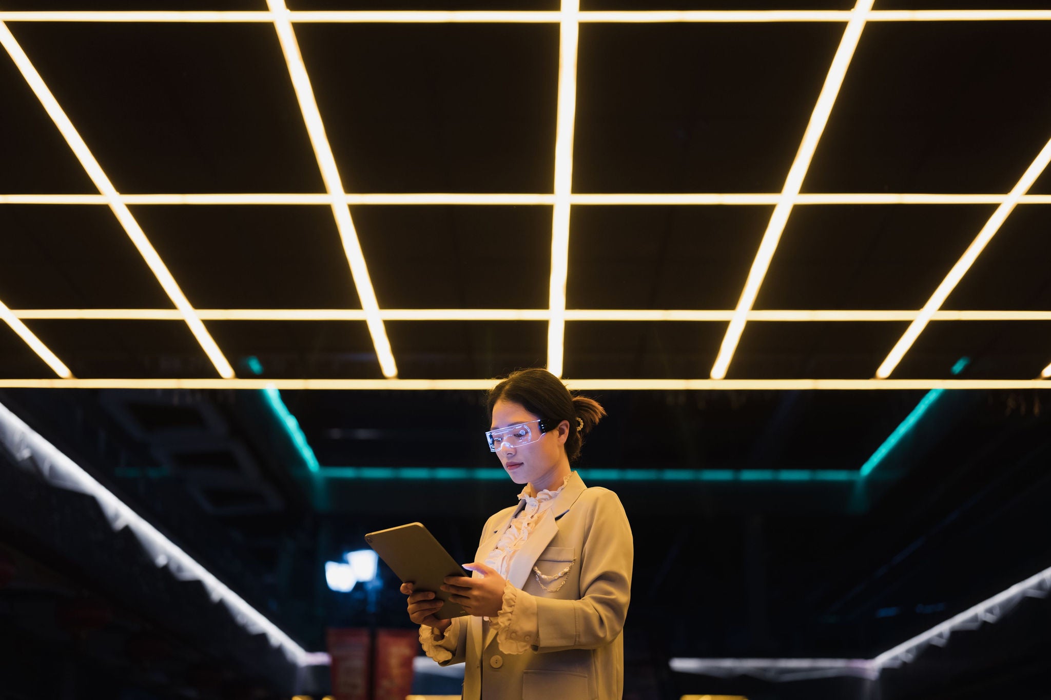 A young Asian business woman wearing glowing smart glasses uses a tablet in a city street at night. Concepts such as augmented reality, artificial intelligence, smart cities, metaverse, post-humanism are expressed.