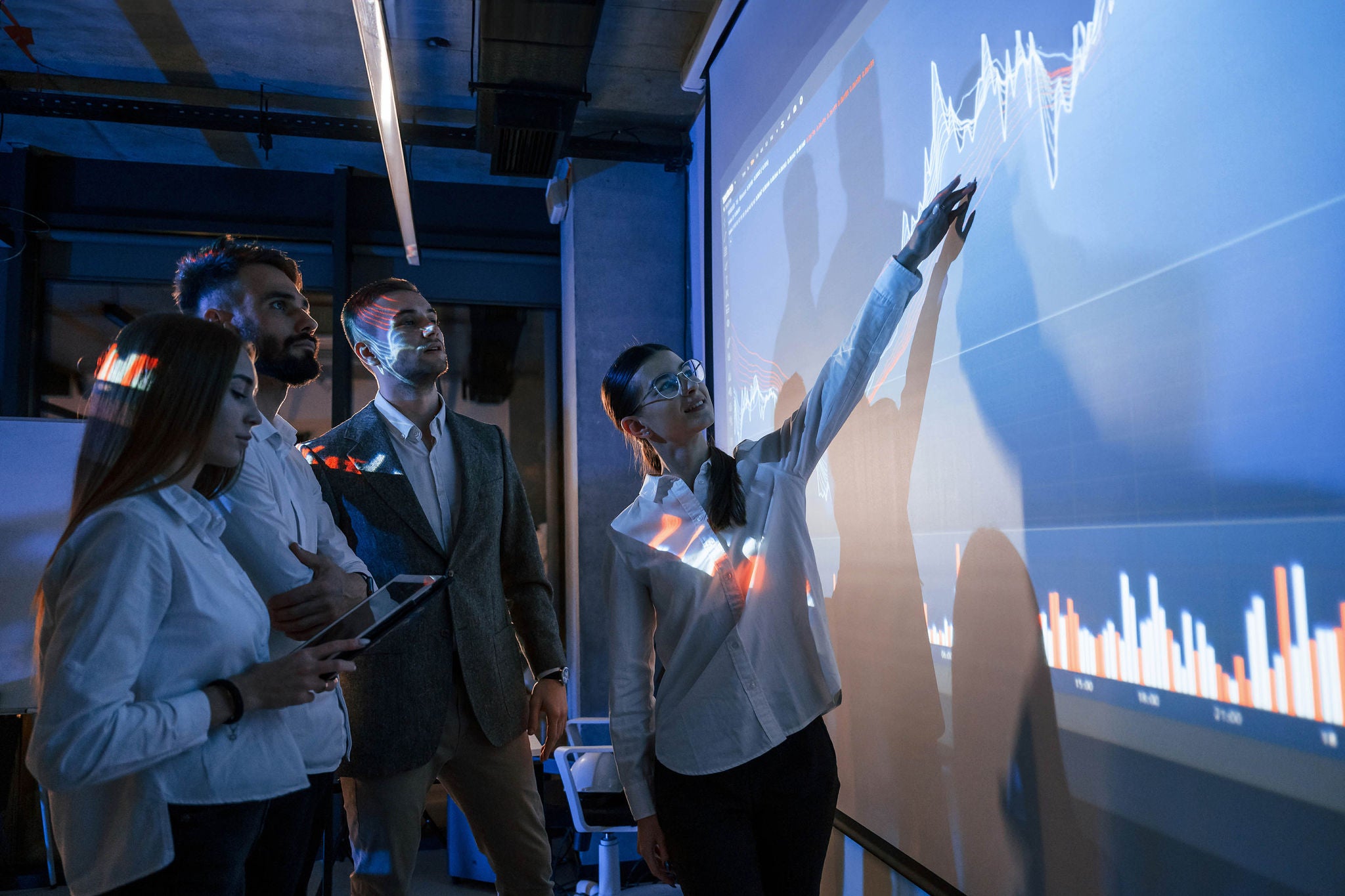 Holding tablet. Male leader talking to employees, showing the plan on the projector in office of stock exchange company.