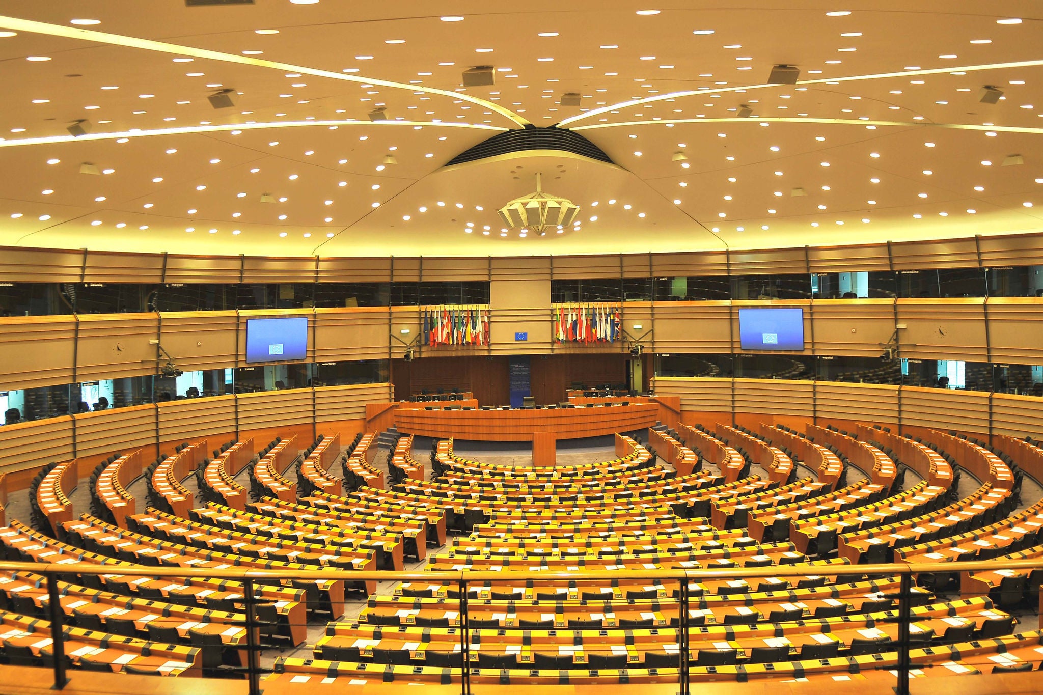 Empty big assembly room of European Parliament in Brussels, Belgium, Europe. Wooden chairs in semi-circle, papers on the banks, lots of boxes for translaiters, European flags and billboards in the background, many lights on the ceiling, fence in front. No people seen. Sandy brown color.