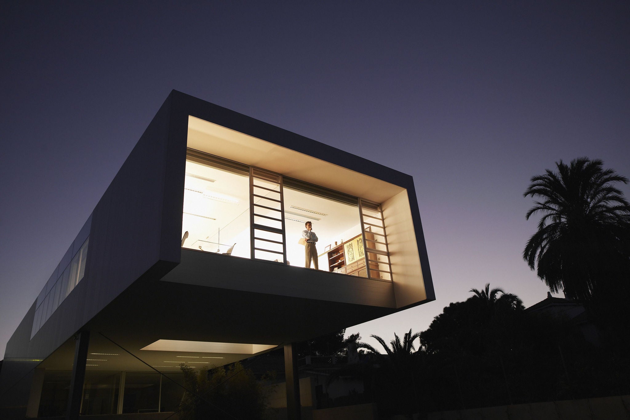  A man thinking by the window staring outside from a modern house with a garden