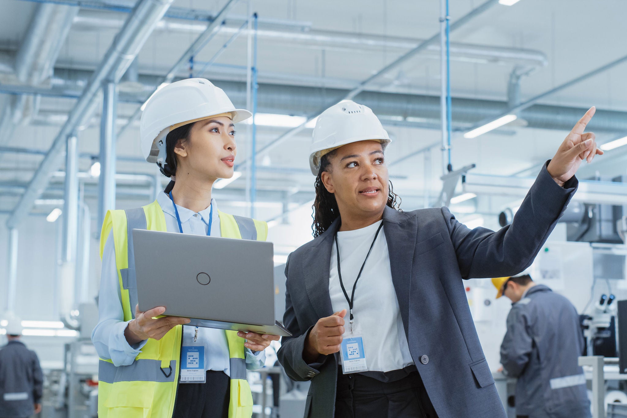 Portrait of Two Female Employees in Hard Hats at Factory. Discussing Job Assignments at Industrial Machine Facility, Using Laptop Computer. Asian Engineer and African American Technician at Work.