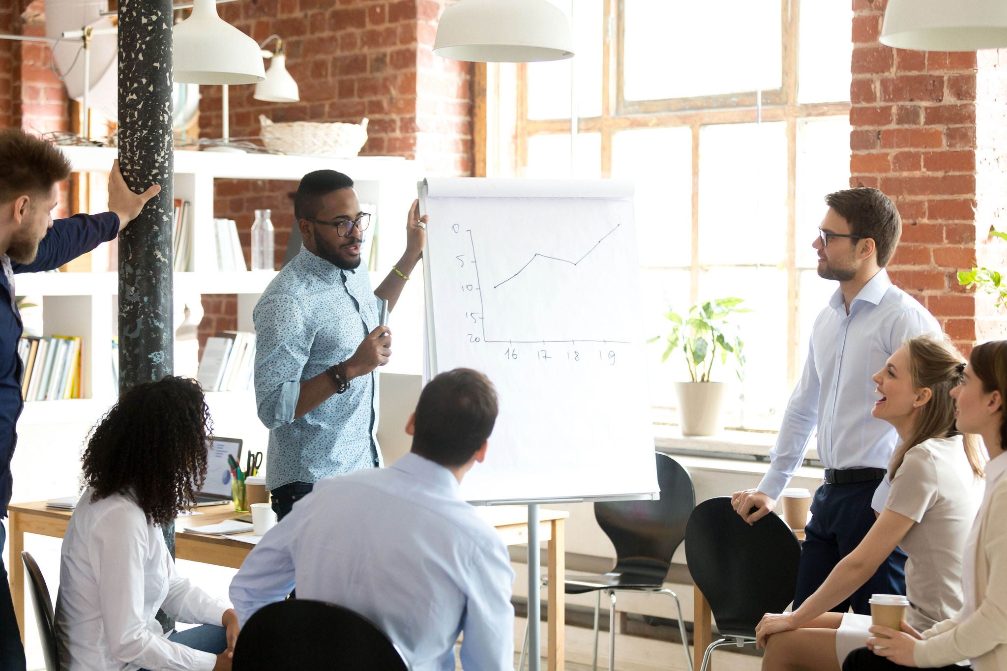 Confident smiling African American business coach speaking presentation to colleagues group at briefing, company meeting, explaining growth of sales graph, reporting about good result, motivating workers