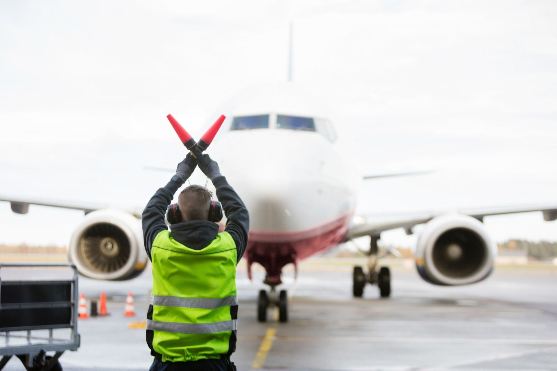 Hombre señalizando el camino en una pista de aterrizaje de un aeropuerto. Frente a él hay un avión.