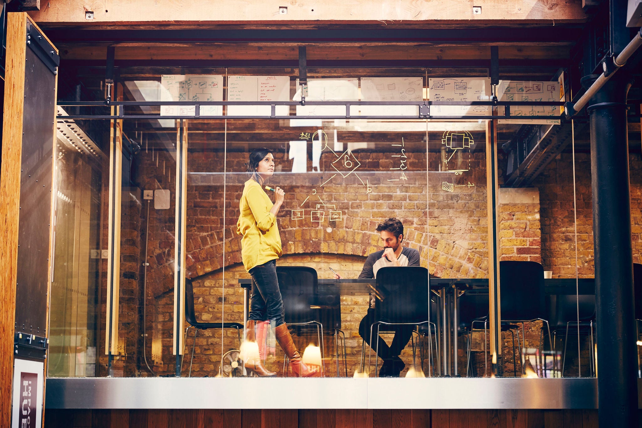 Two creative professionals work on a project, mind mapping ideas onto glass window, in modern office space.