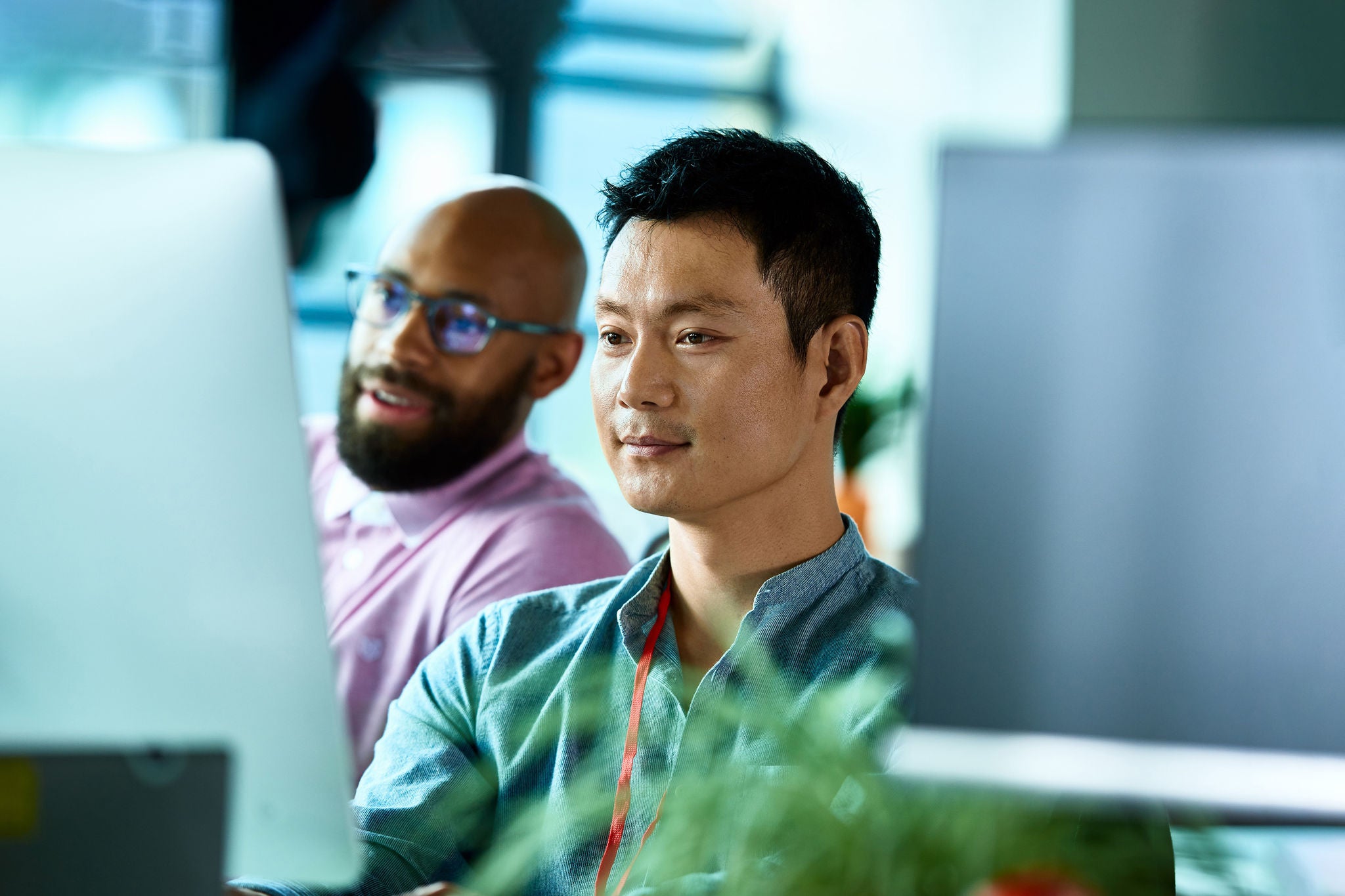 Two male co-workers looking at computer screen and concentrating, mid adult Asian man wearing lanyard, freelance worker